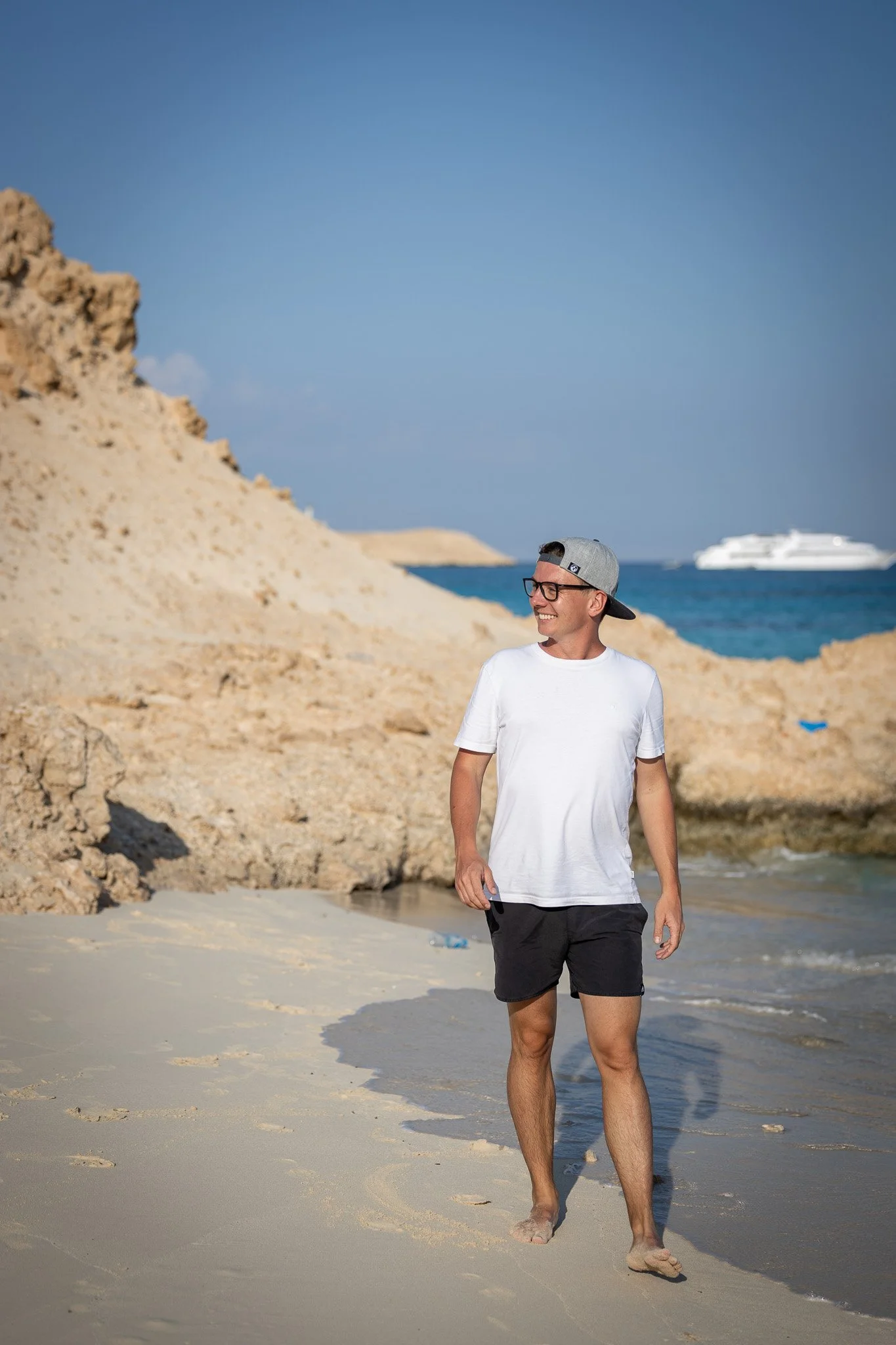 Sebastian Schmidt am Strand mit Sand, Felsen, Meer und einem Kreuzfahrtschiff im Hintergrund