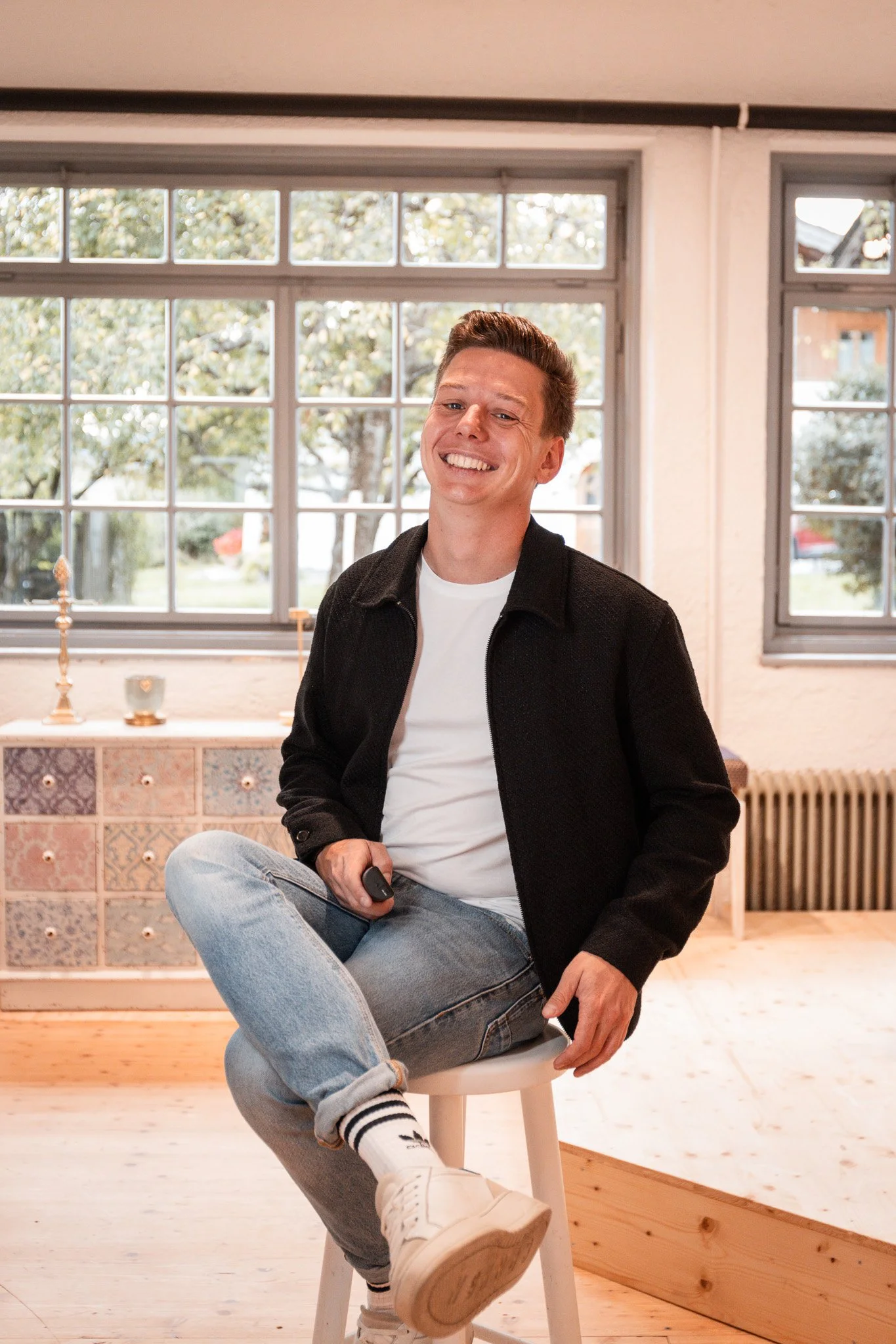 Sebastian Schmidt sitzt auf einem Hocker in einem hellen Raum, lächelt freundlich und hält eine Fernbedienung in der Hand, im Hintergrund große Fenster mit Blick nach draußen.