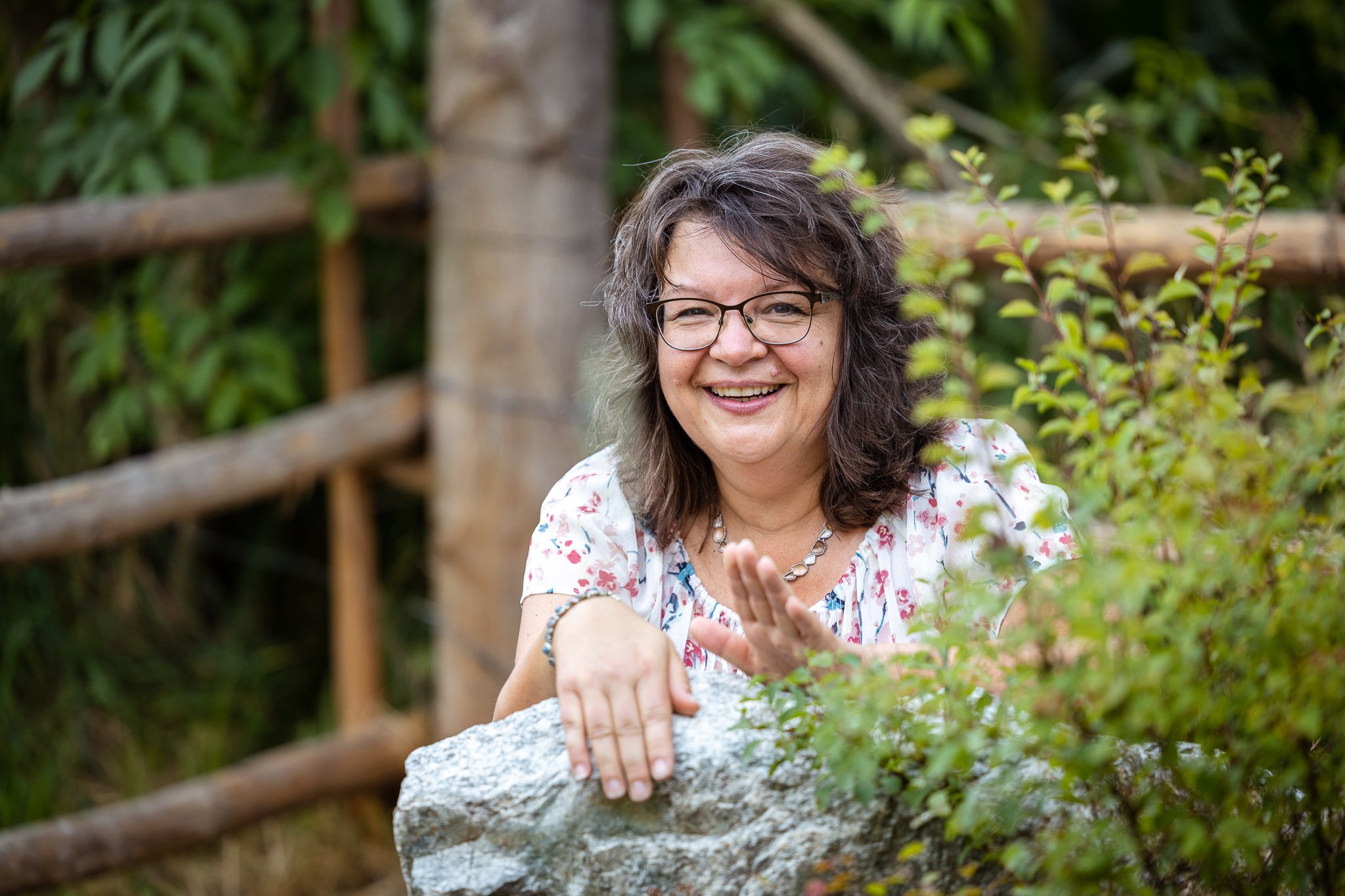 Lächelnde Frau mit Brille und Schmuck, die auf einen Felsen lehnt, im Freien mit Holzzaun im Hintergrund.