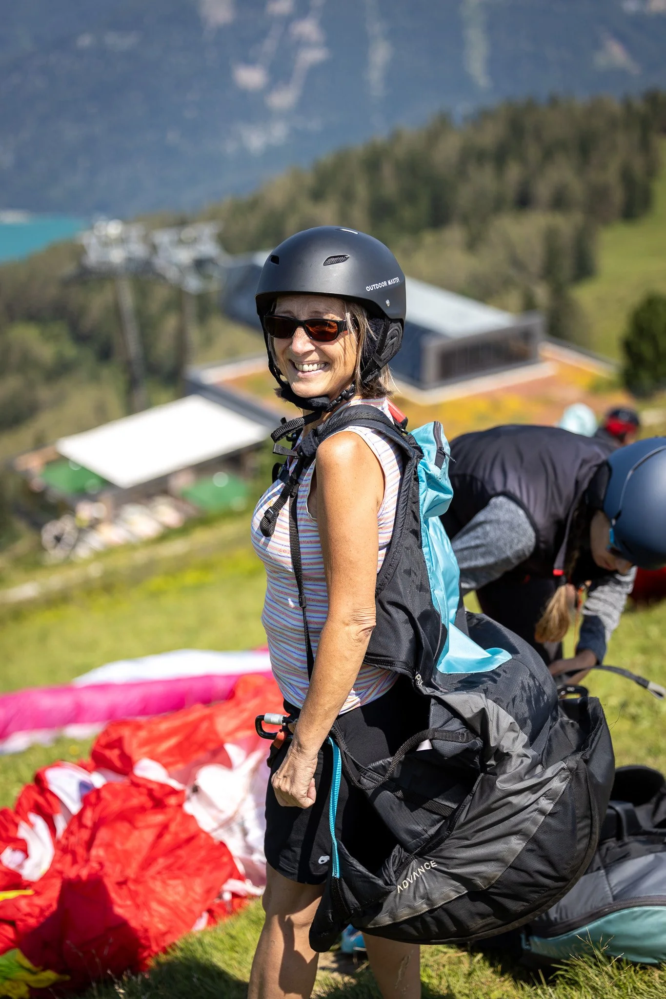 Eine lachende Frau mit Helm und Sonnenbrille steht in einem alpinen Gelände, umgeben von Berglandschaft und einem Skilift im Hintergrund, während sie mit einem Rucksack für einen Paragliding-Ausflug vorbereitet.