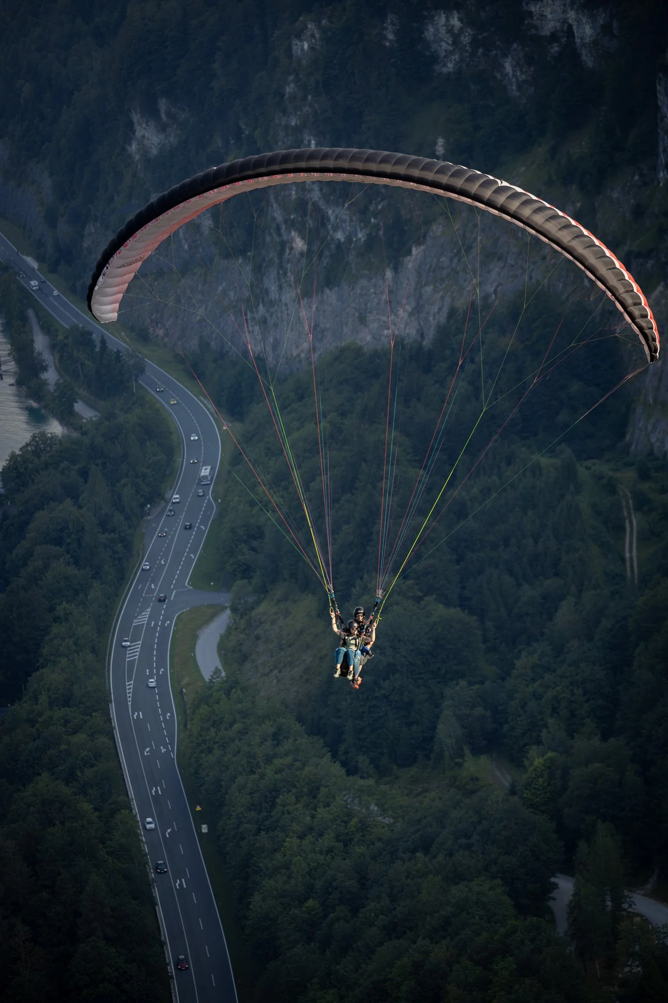 Zwei Menschen beim Paragliding über einer kurvigen Straße, umgeben von grünen Wäldern und Bergen.