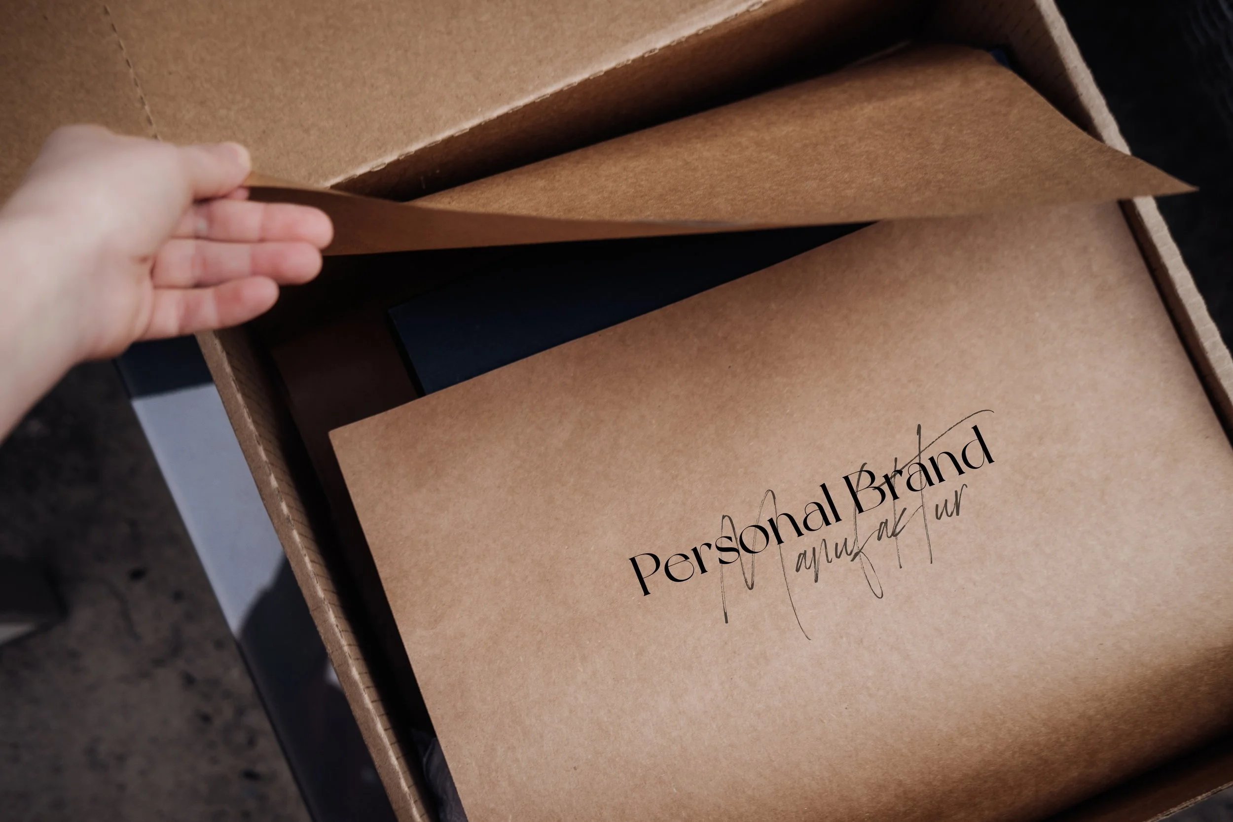 Eine Hand hebt eine braune Papierfolie aus einer offenen Kartonschachtel. Darin befindet sich ein Dokument mit der Überschrift 'Personal Brand audit'.