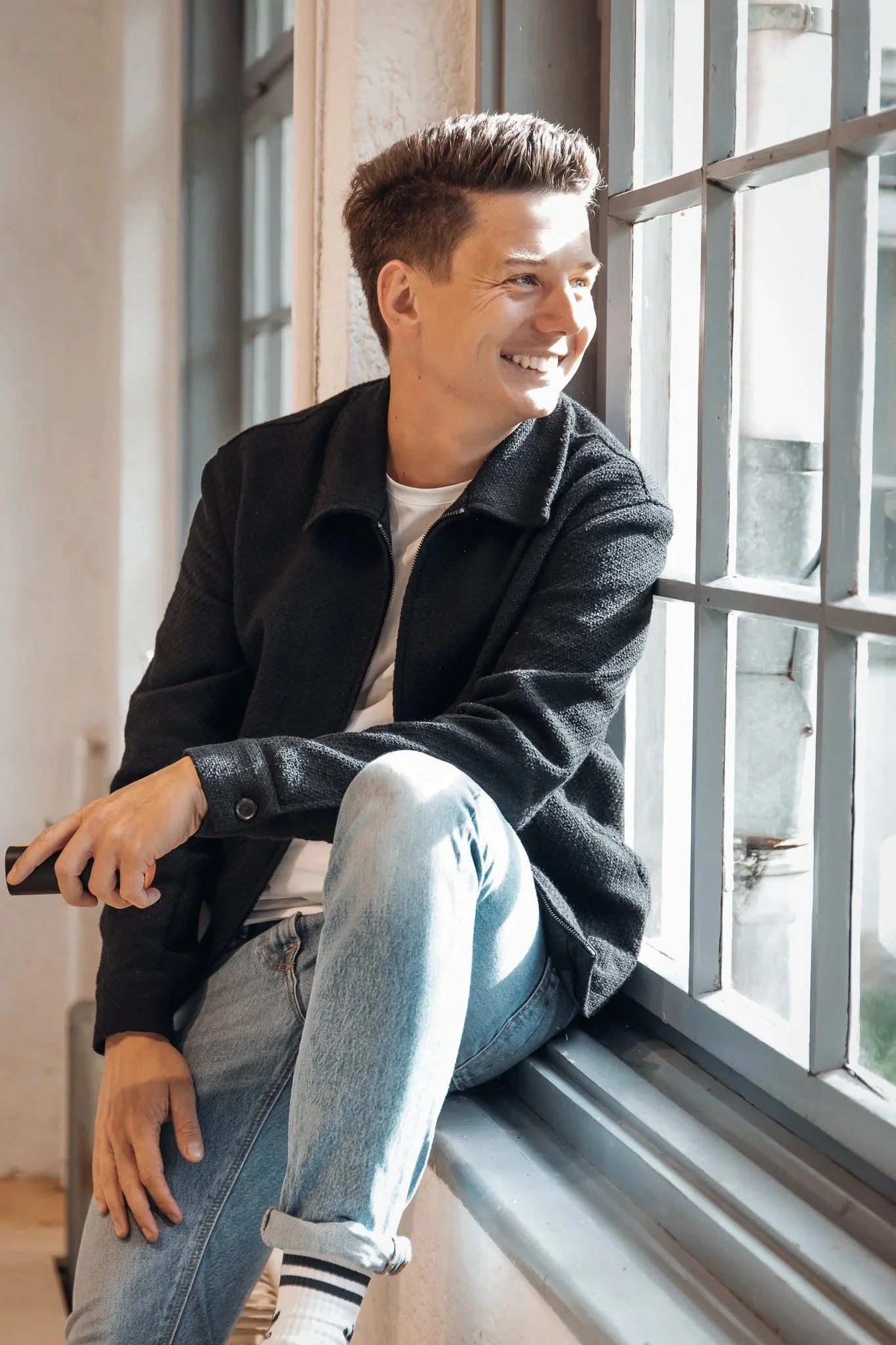 Sebastian Schmidt sitzt an einem Fenster, trägt eine schwarze Jacke, ein weißes T-Shirt und Jeans, im Sonnenlicht.