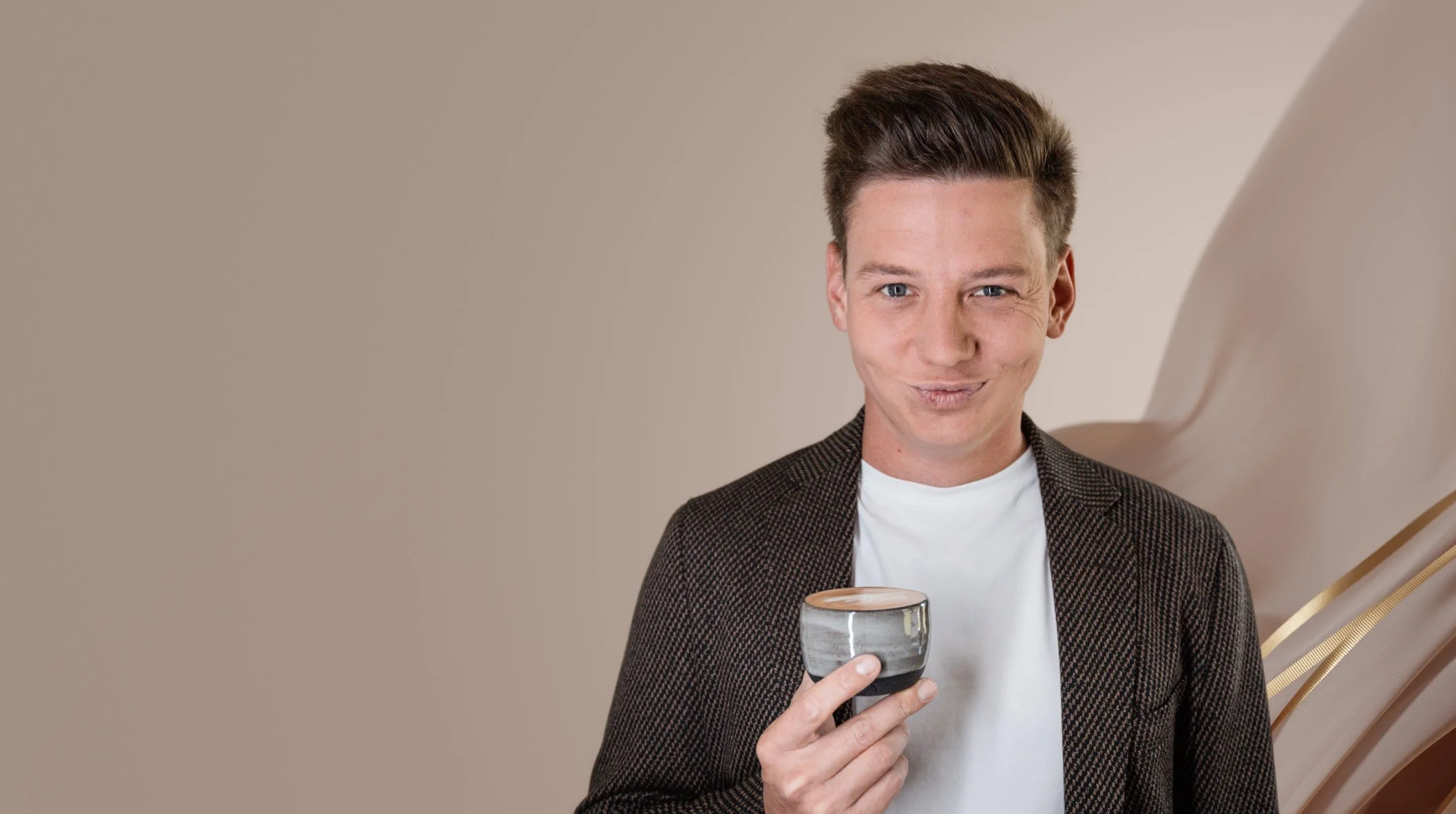 Sebastian Schmidt mit kurzen braunen Haaren, trägt einen schwarzen Hemd mit weißen Punkten und hält eine Tasse Kaffee in der Hand. Er lächelt verschmitzt und steht vor einem beige-grauen Hintergrund.