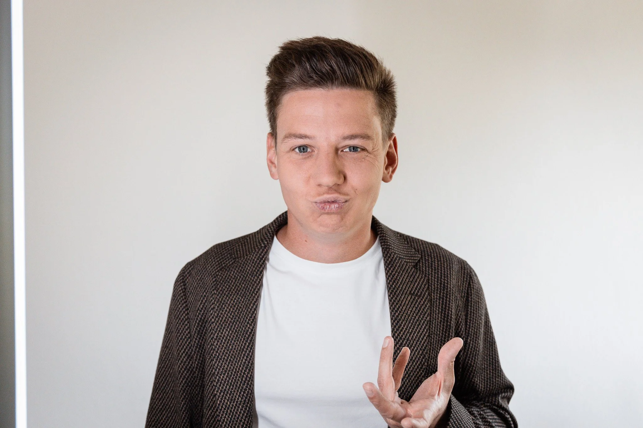Junger Mann macht eine tanzende Geste und köpft die Lippen, trägt ein weißes T-Shirt und eine braune Jacke, steht vor einem hellen Hintergrund.