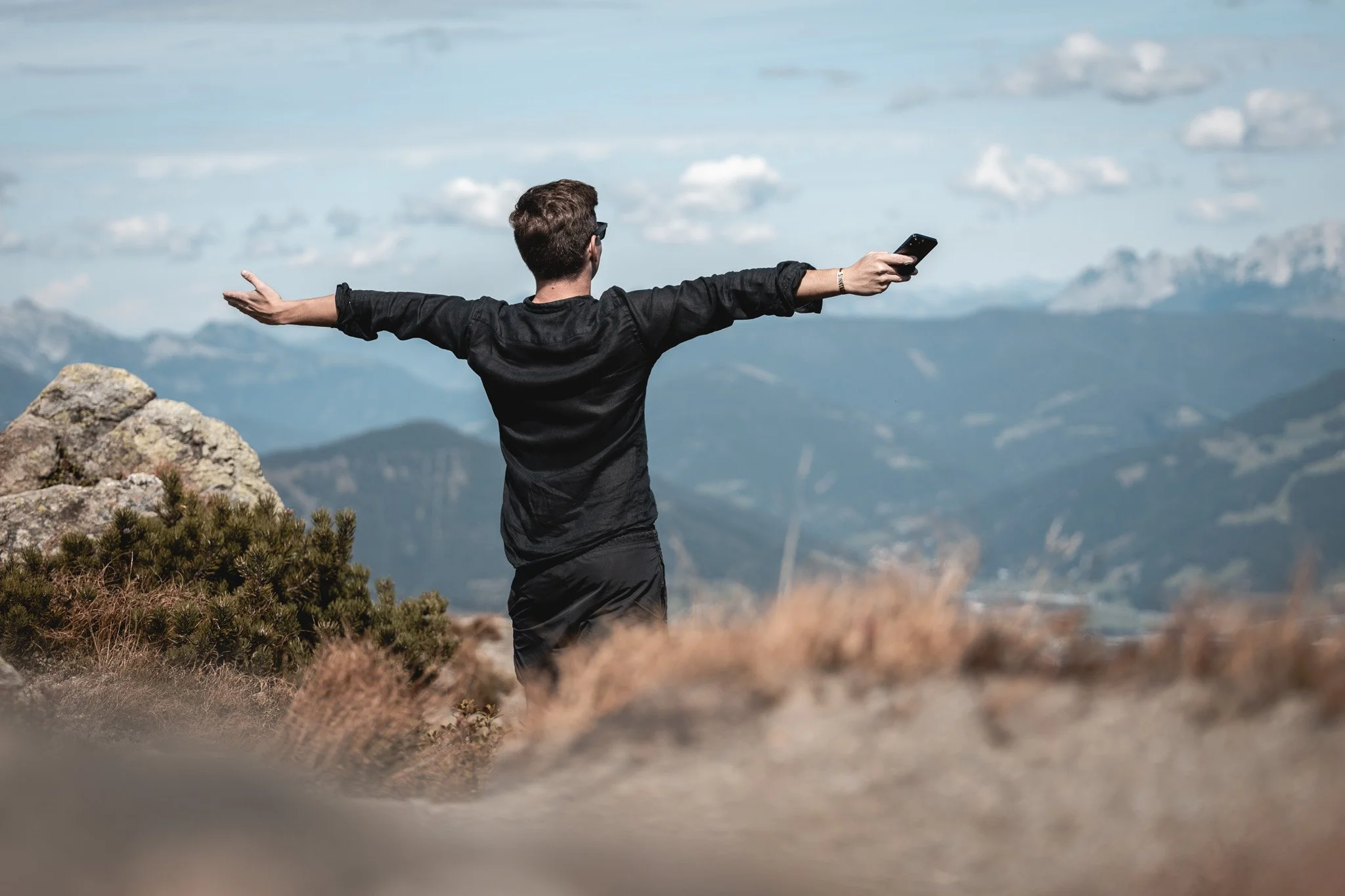 Sebastian Schmidt steht auf einem Berg mit offenem Blick, breiten Armen und hält ein Smartphone in der rechten Hand, um die Aussicht zu genießen.