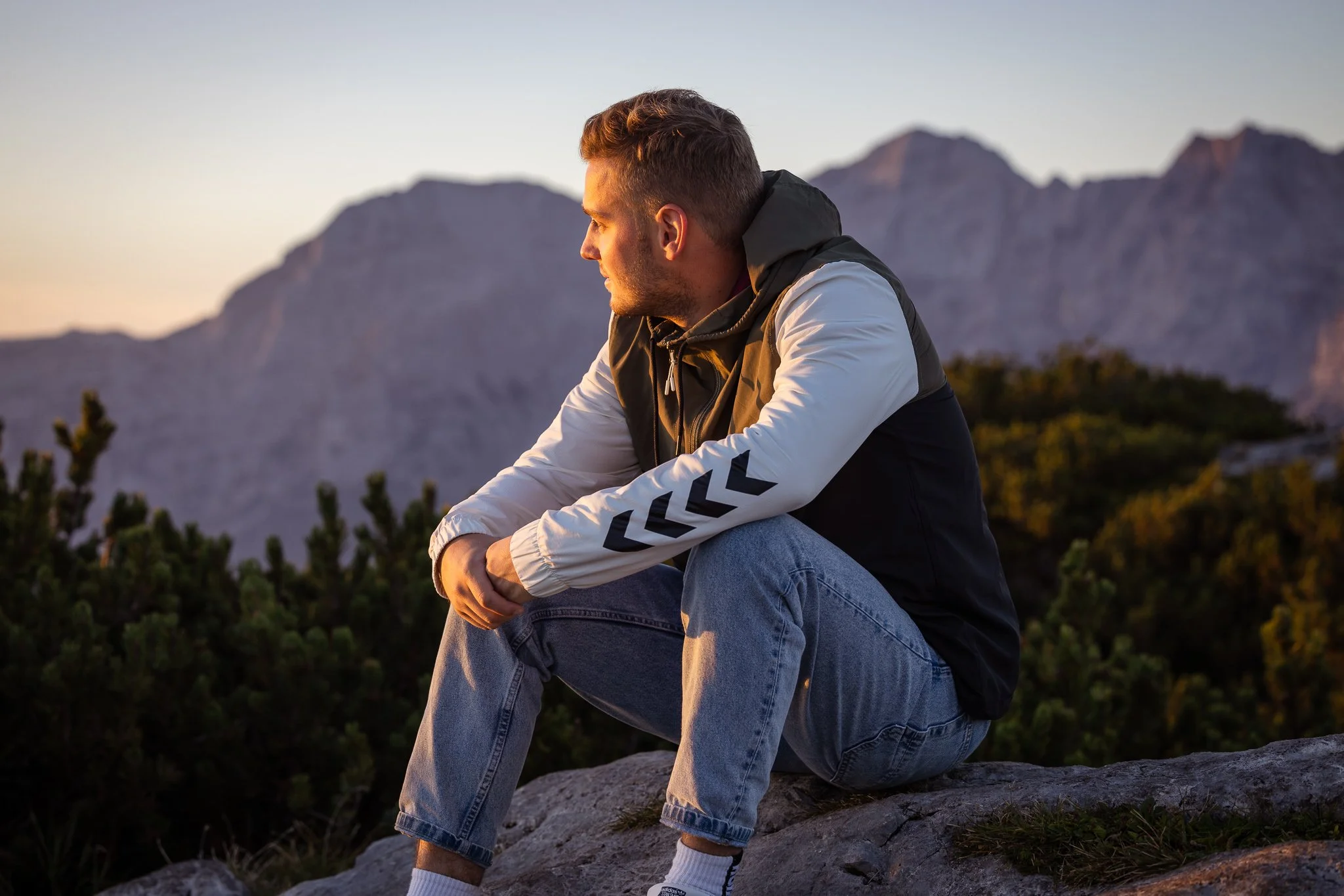 Junge sitzt auf einem Felsen in den Bergen bei Sonnenuntergang, trägt eine schwarze und weiße Jacke und blaue Jeans.
