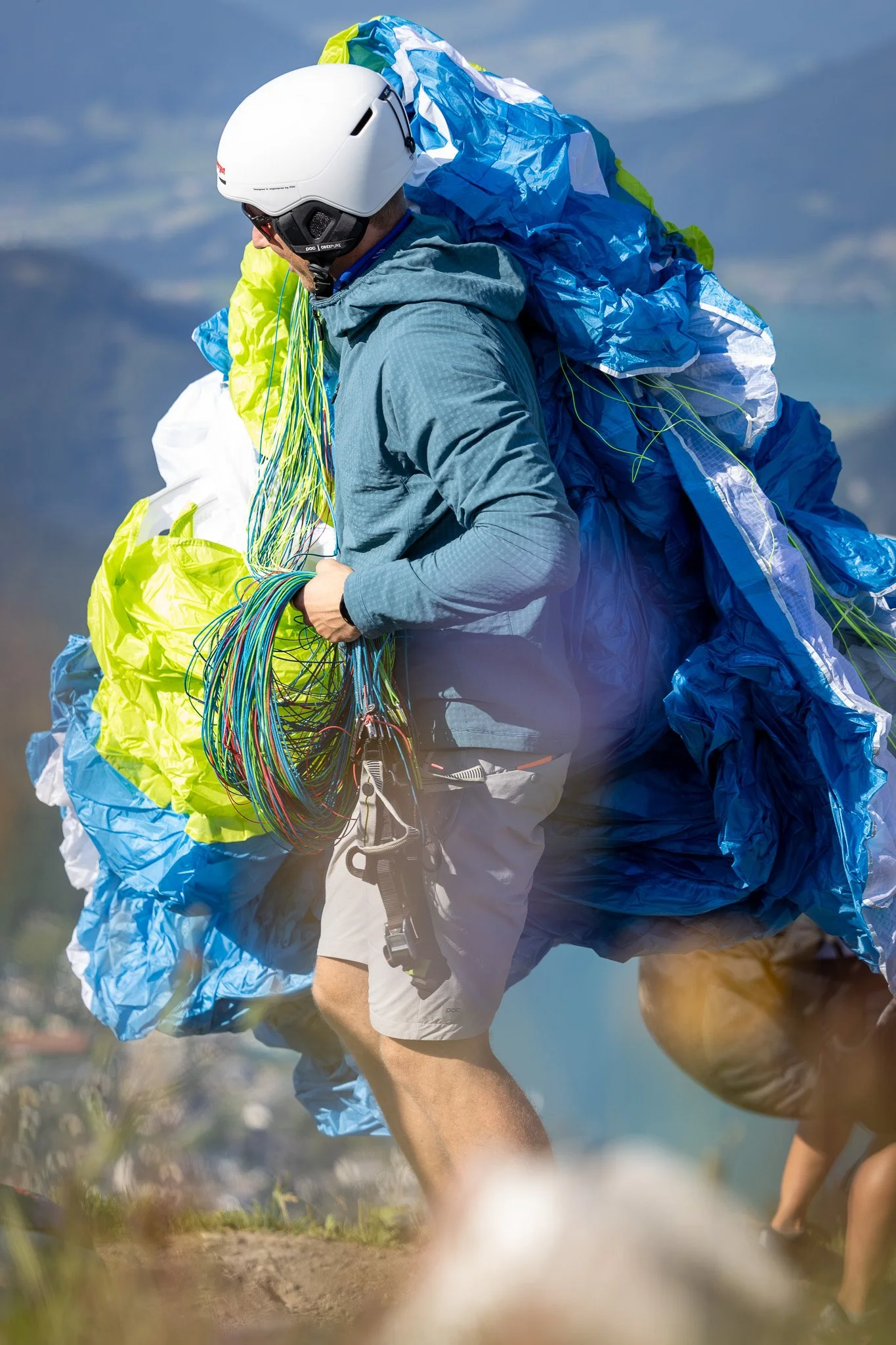Mann beim Paragliding-Start, trägt Schutzhelm und Jacke, hält viele farbige Gleitschirmleinen, mit einem türkisfarbenen, blauen und grünen Gleitschirm auf einem Berg.