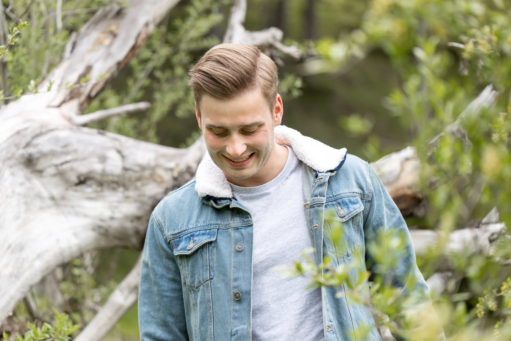 Junge Mann steht im Wald vor umgestürztem Baum, trägt Jeansjacke und graues T-Shirt, lächelt und schaut nach unten.
