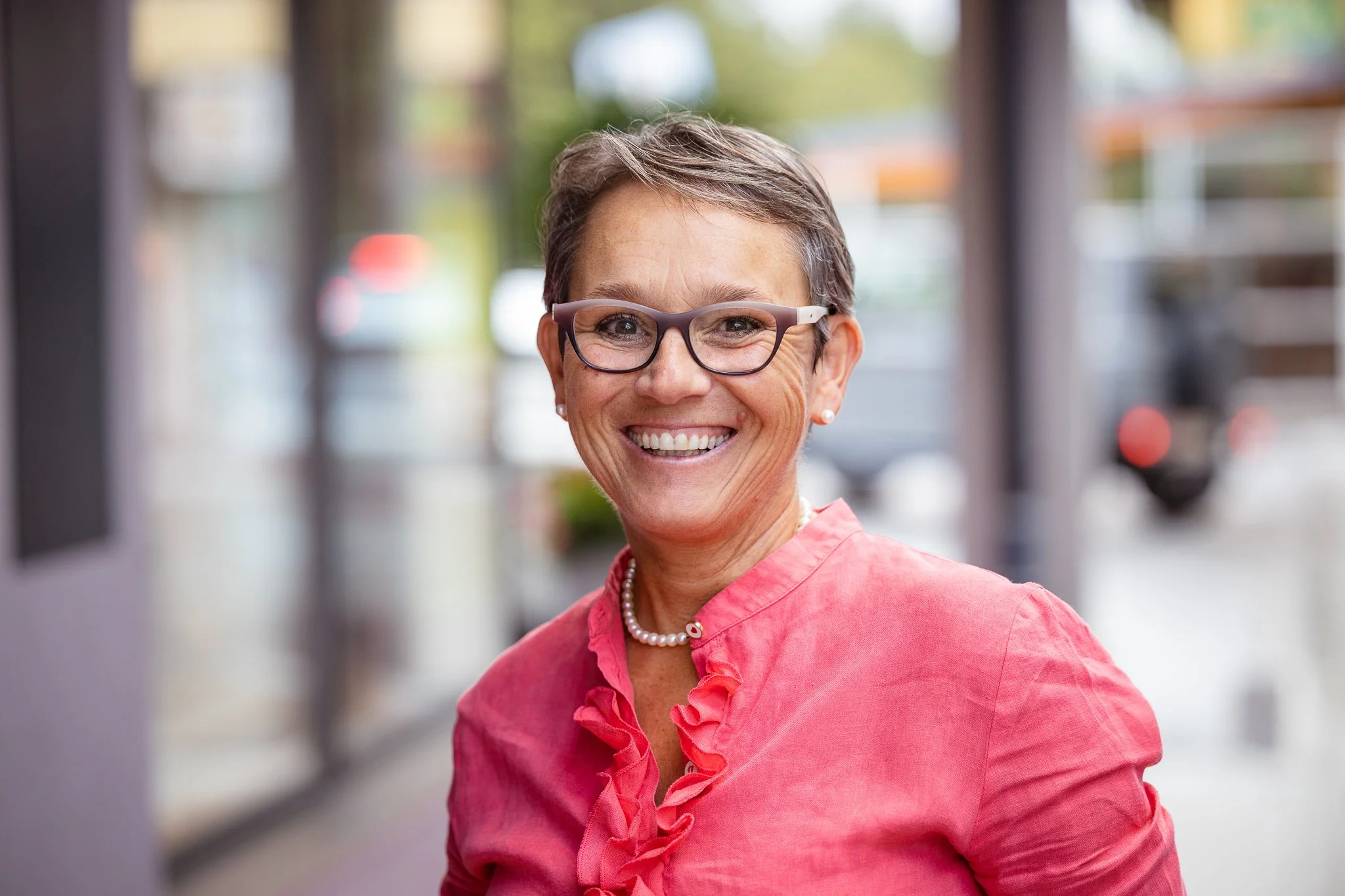 Ein lächelnde ältere Frau mit Kurzhaarschnitt, Brille, Perlenkette und rotem Bluse vor einem Gebäude mit Glasfenstern.