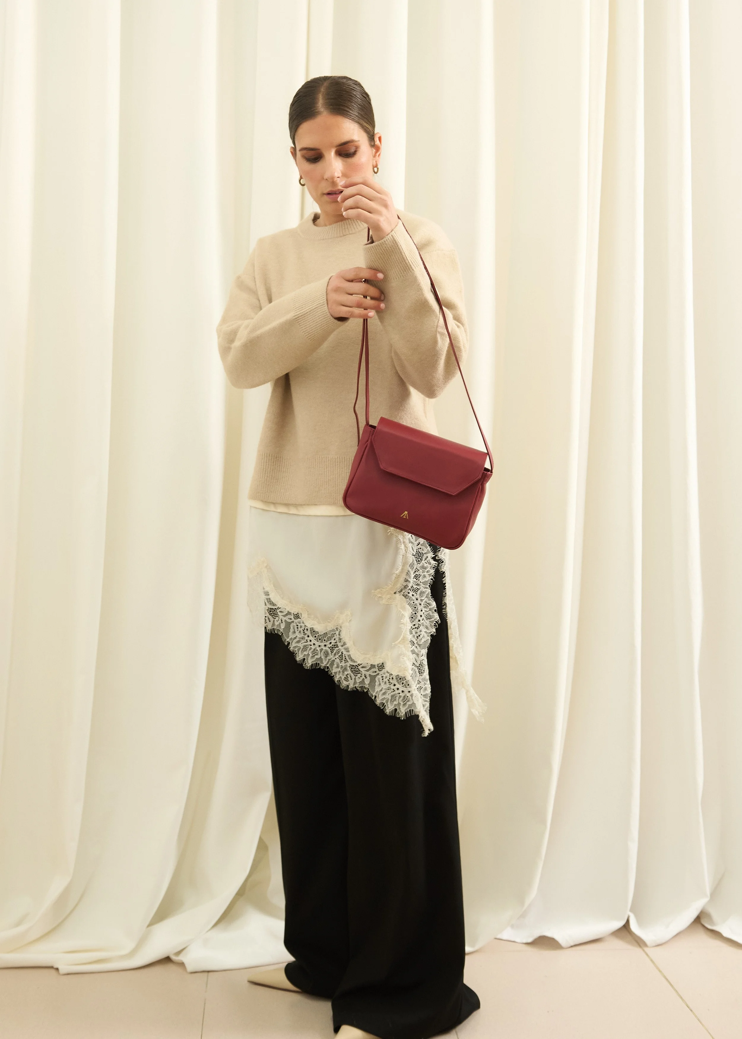 Mujer con cabello castaño oscuro, usando suéter beige, falda larga negra, y una bufanda de encaje blanca, sostiene un bolso burdeos, en un fondo de cortinas blancas.