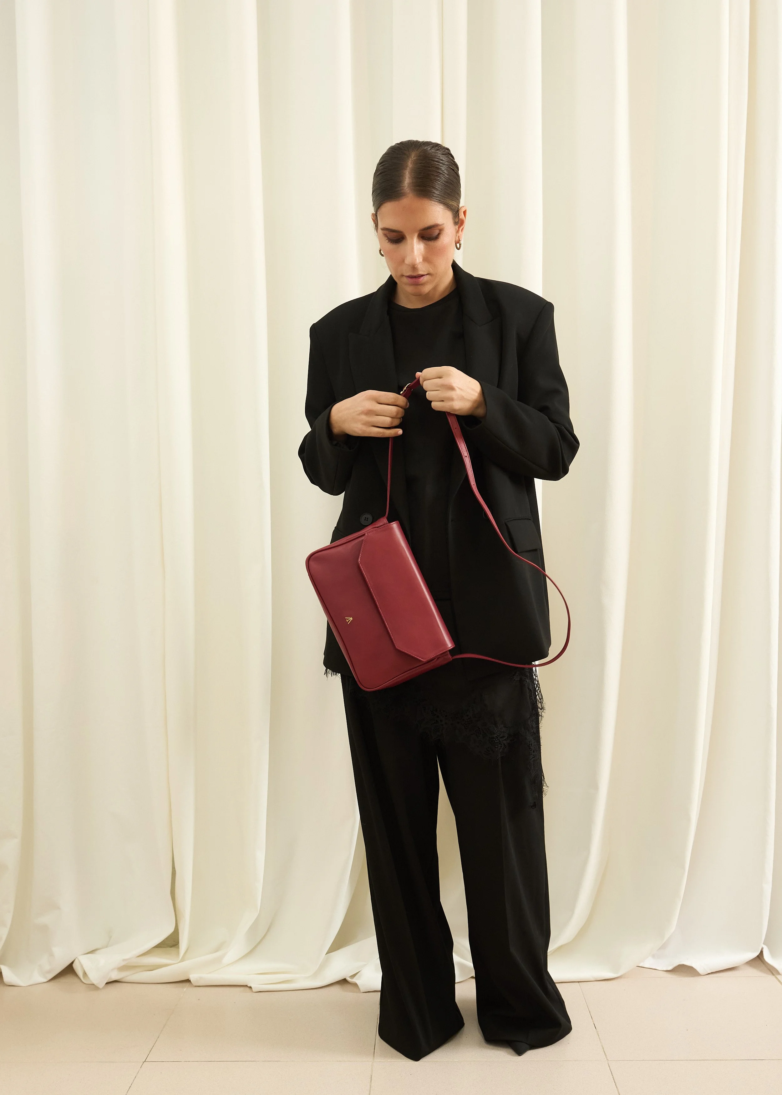 Mujer con ropa negra y bolso rojo, ajustando la correa del bolso en un fondo de cortinas blancas.