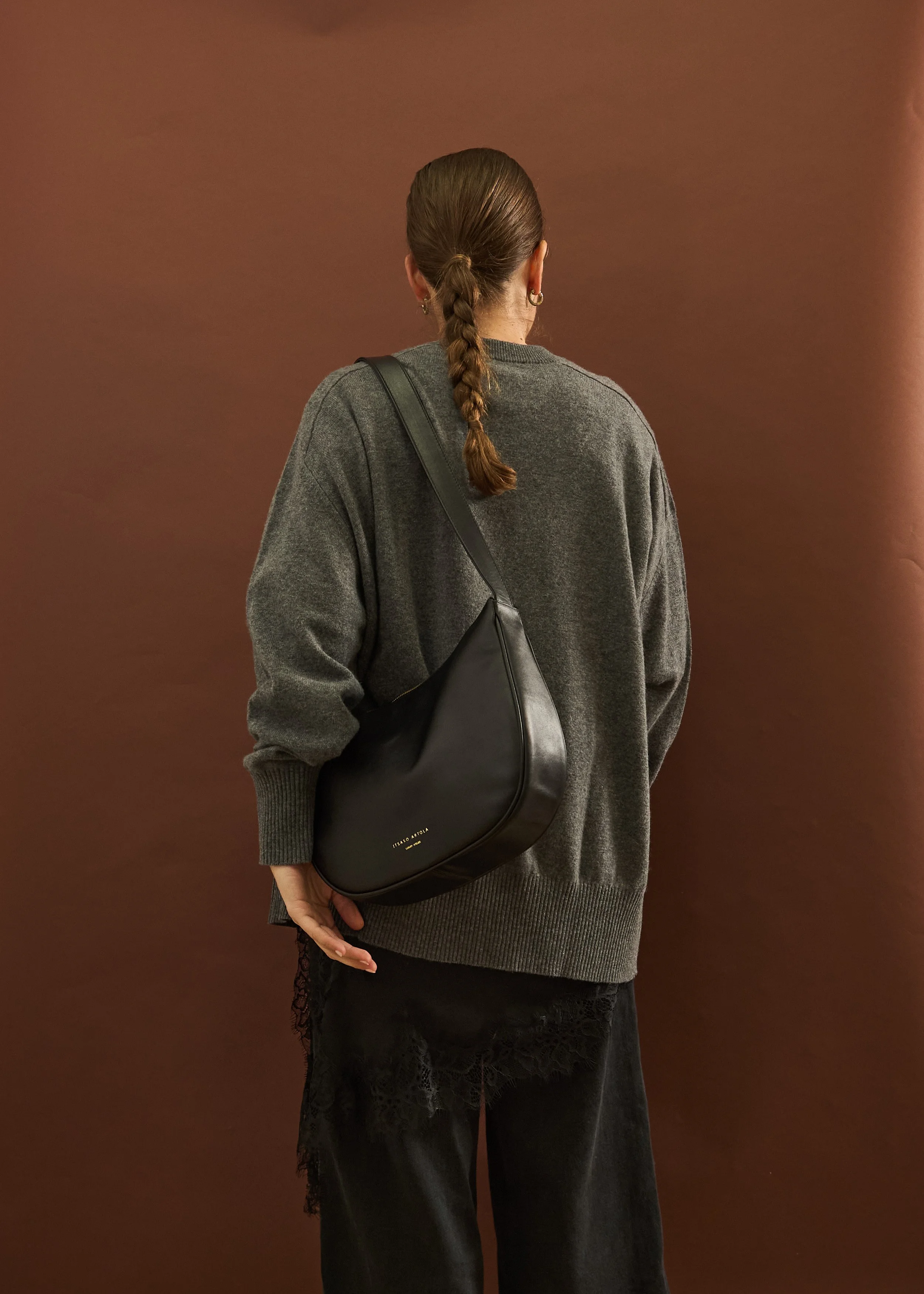 Mujer con cabello rizado en coleta, vista de espaldas, lleva suéter gris oscuro y pantalones negros con encaje, con bolso negro sobre su hombro, frente a fondo marrón.