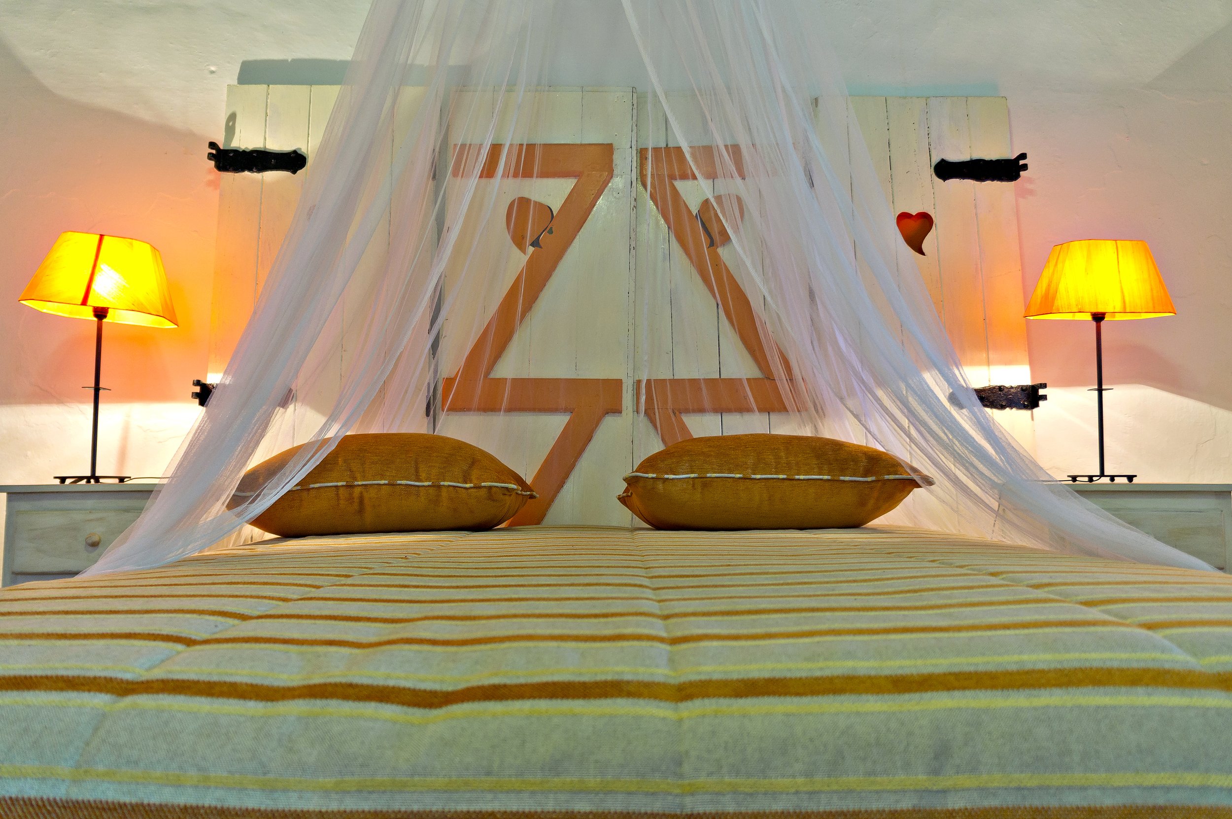 Bedroom with striped bedding, two yellow cushions, decorative headboard, lamps on nightstands, and white canopy drapes.