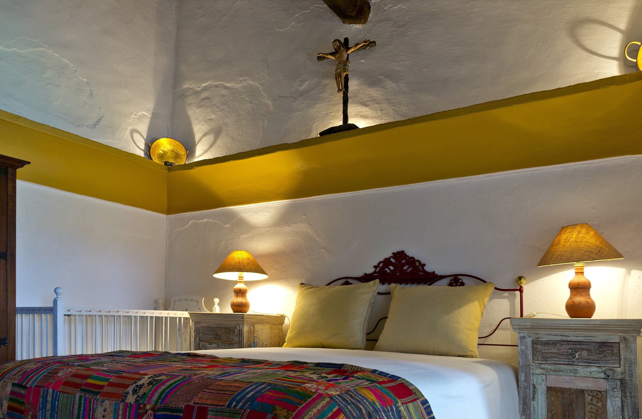 Cozy bedroom with colorful quilt, cross on wall, two wooden bedside tables with lamps, and neutral pillows on the bed.