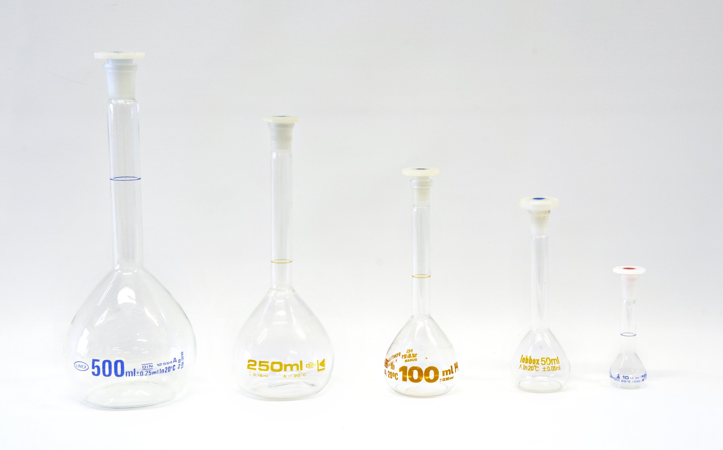 Five different sized glass volumetric flasks with stoppers, labeled with their capacities: 500ml, 250ml, 100ml, 50ml, and 10ml, aligned in descending order of size.