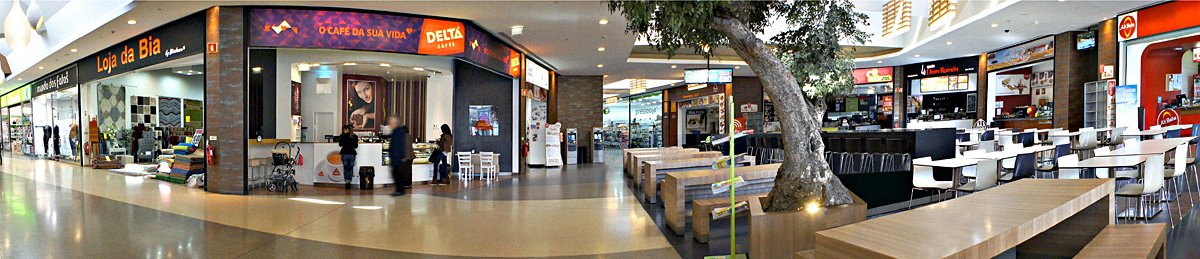 Panoramic view of a shopping mall food court with various stores and eateries, including a coffee shop and retail store, surrounded by seating areas and a tree.