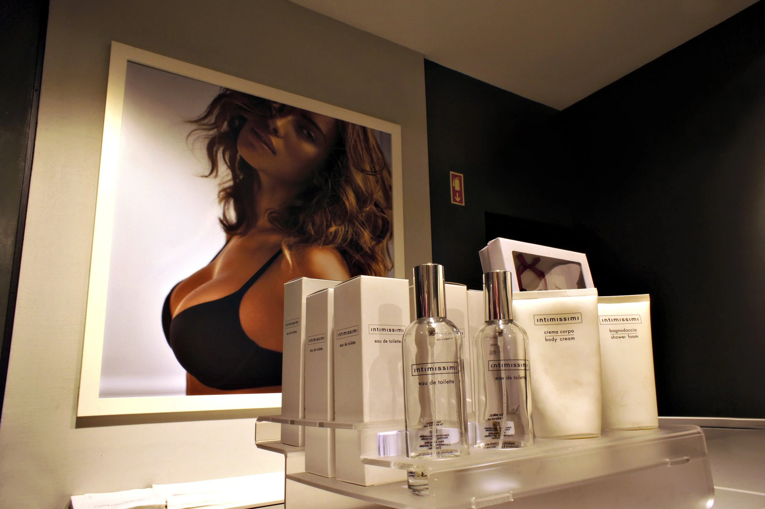 Store display featuring Intimissimi perfume, body cream, and shower foam with a large promotional poster of a woman in a black bra in the background.