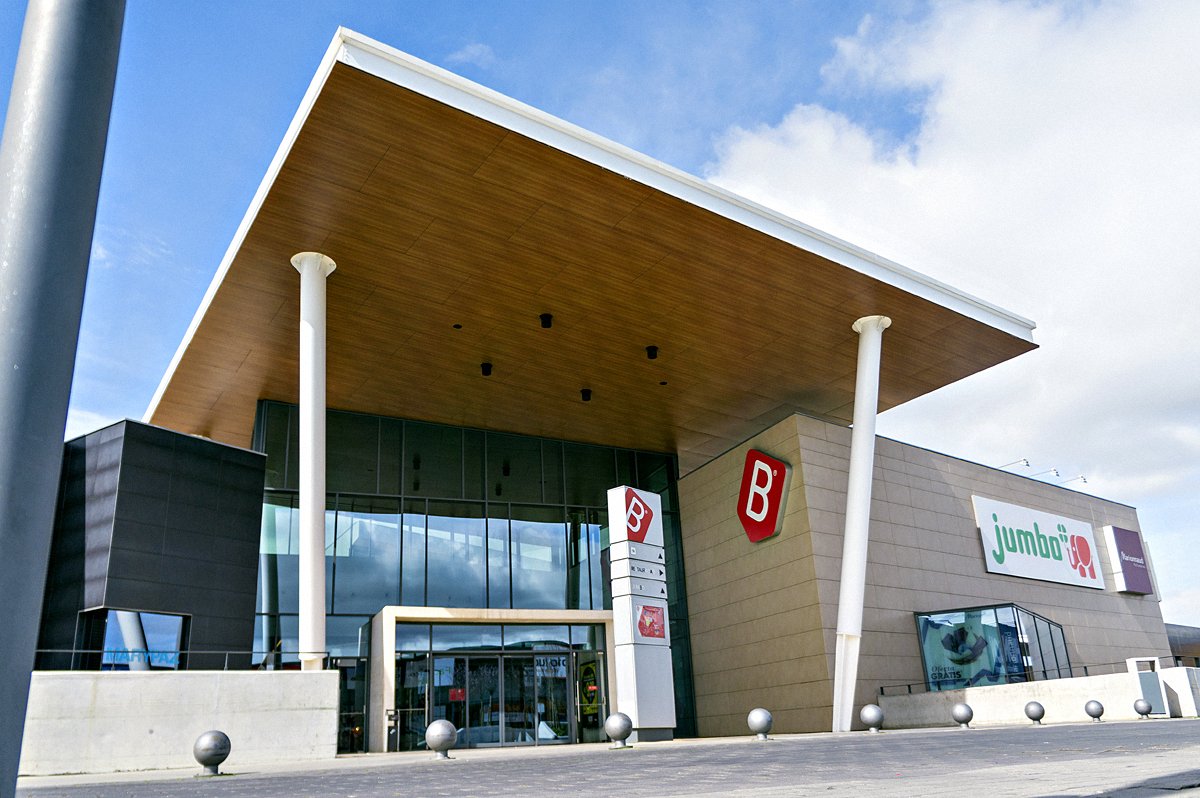 Exterior view of a large modern building with a slanted roof, featuring the logos "B" and "Jumbo," set against a blue sky with clouds.