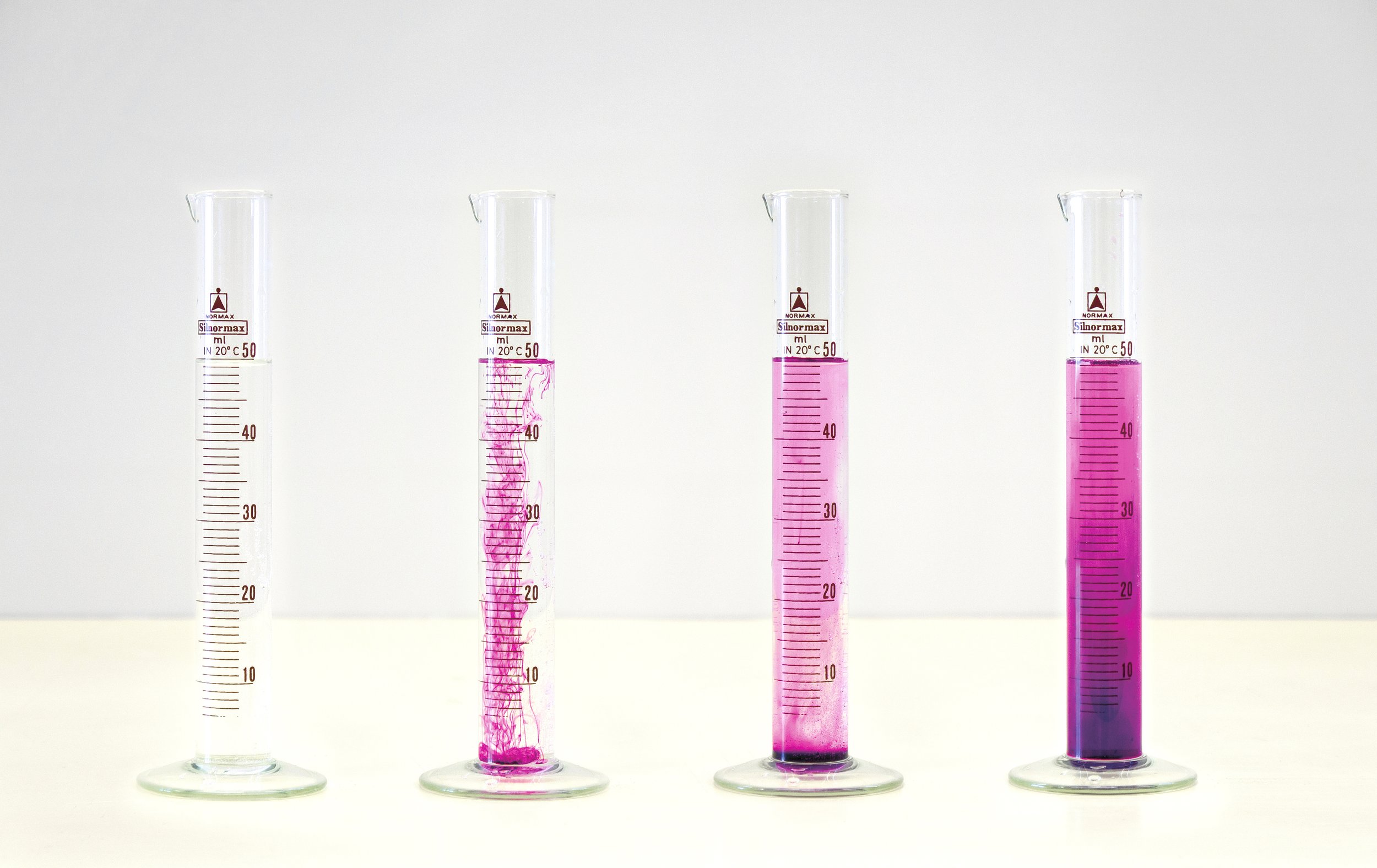 Four graduated cylinders on a table, each containing a fluid transitioning from clear to pink.