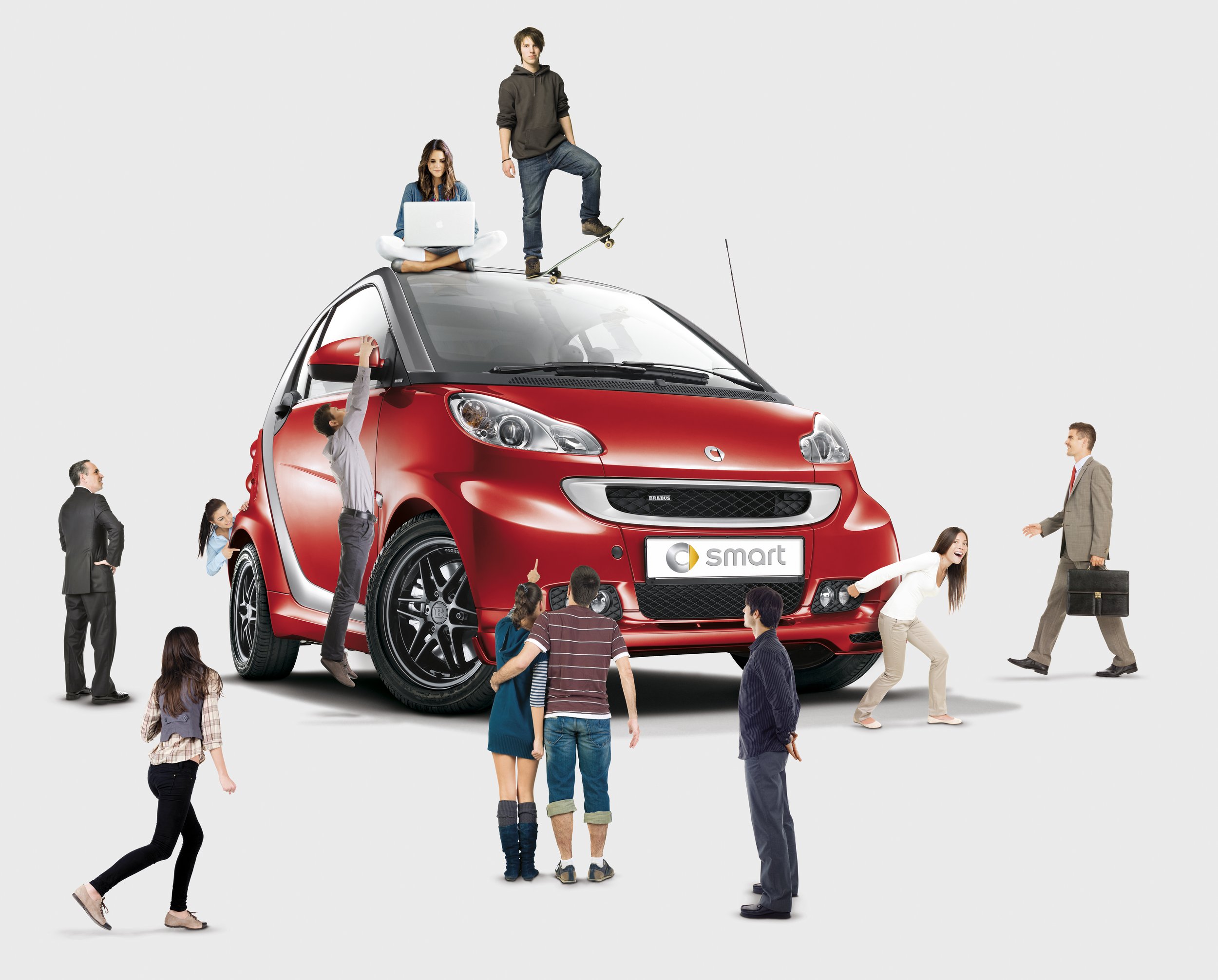 A group of people surround and interact with a red Smart car. A person with a laptop sits on the roof, while others point and look at the car. Some people appear to be examining the vehicle, with one person climbing up to reach the top.
