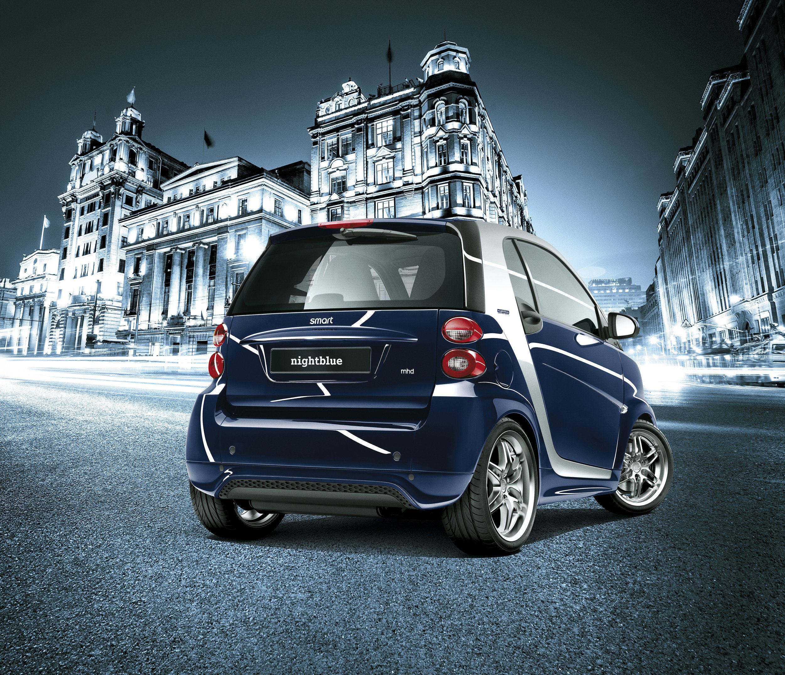 Blue Smart car parked on a city street at night with historic buildings and dynamic lighting in the background.