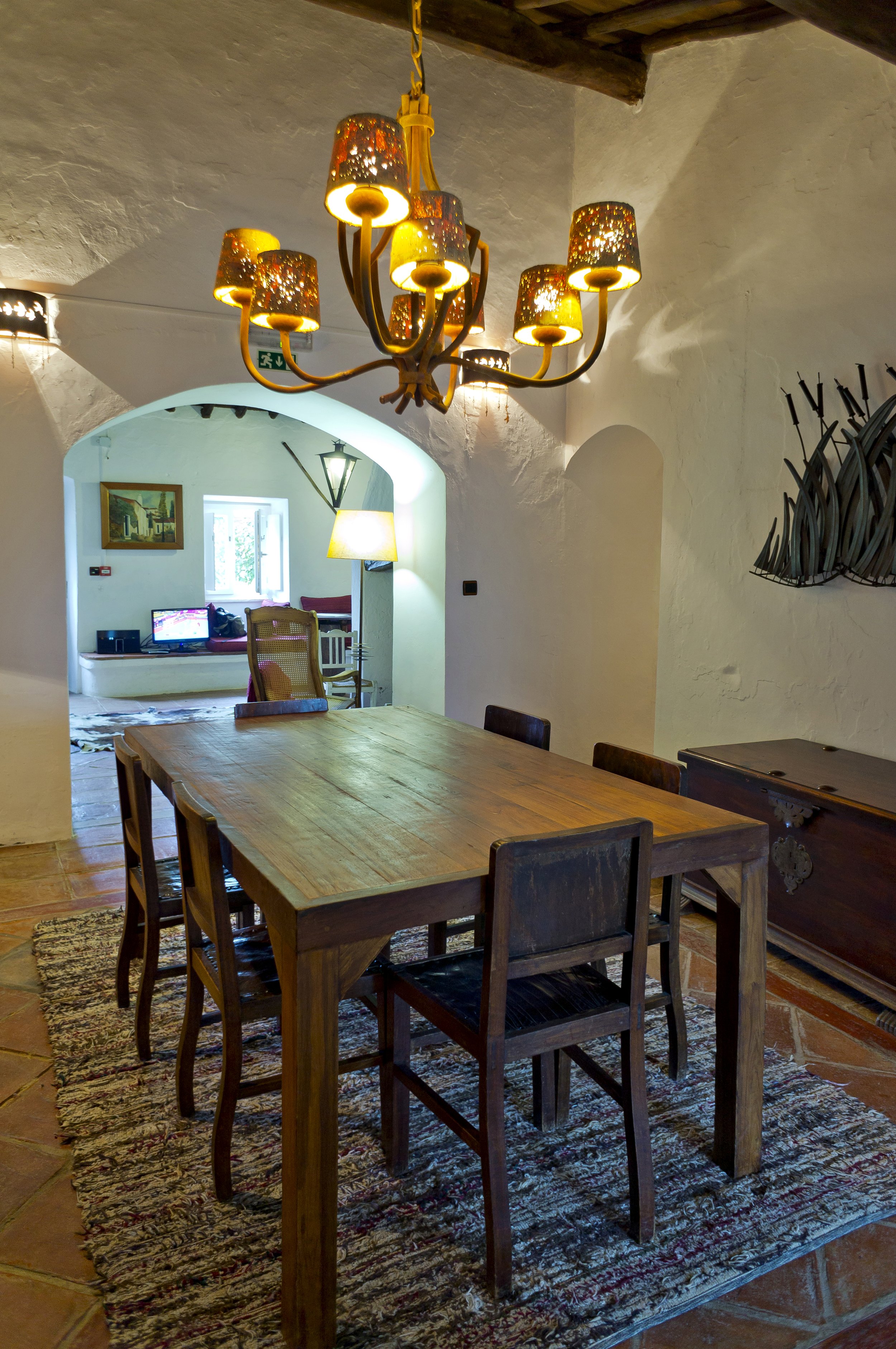 Interior of a rustic dining room with a wooden table, chairs, and a chandelier. The room is decorated with a wall-mounted sculpture and has an adjacent cozy sitting area with a window.