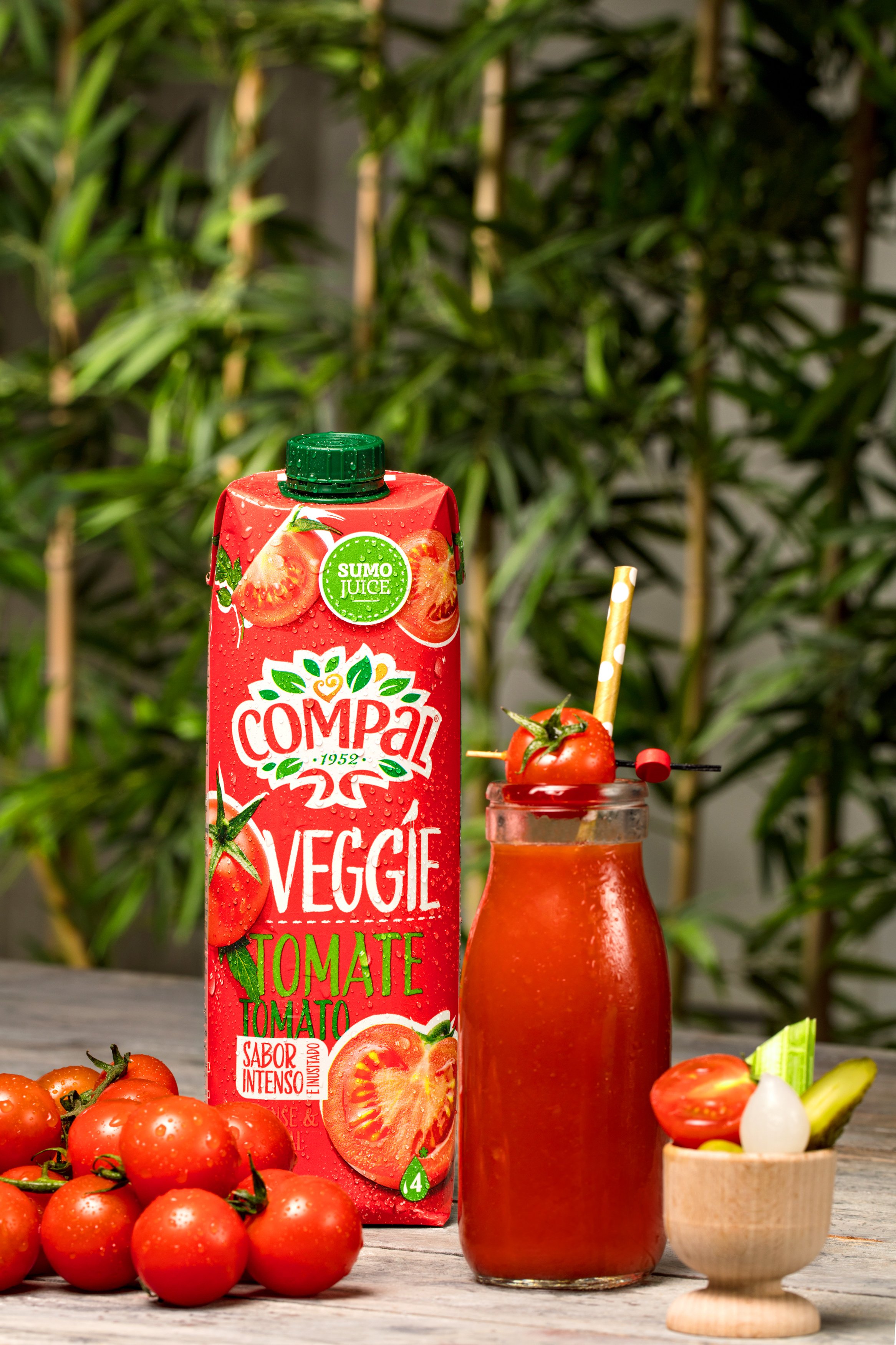 Compal tomato juice carton and glass with fresh tomatoes, bamboo background, and vegetable skewer garnish.