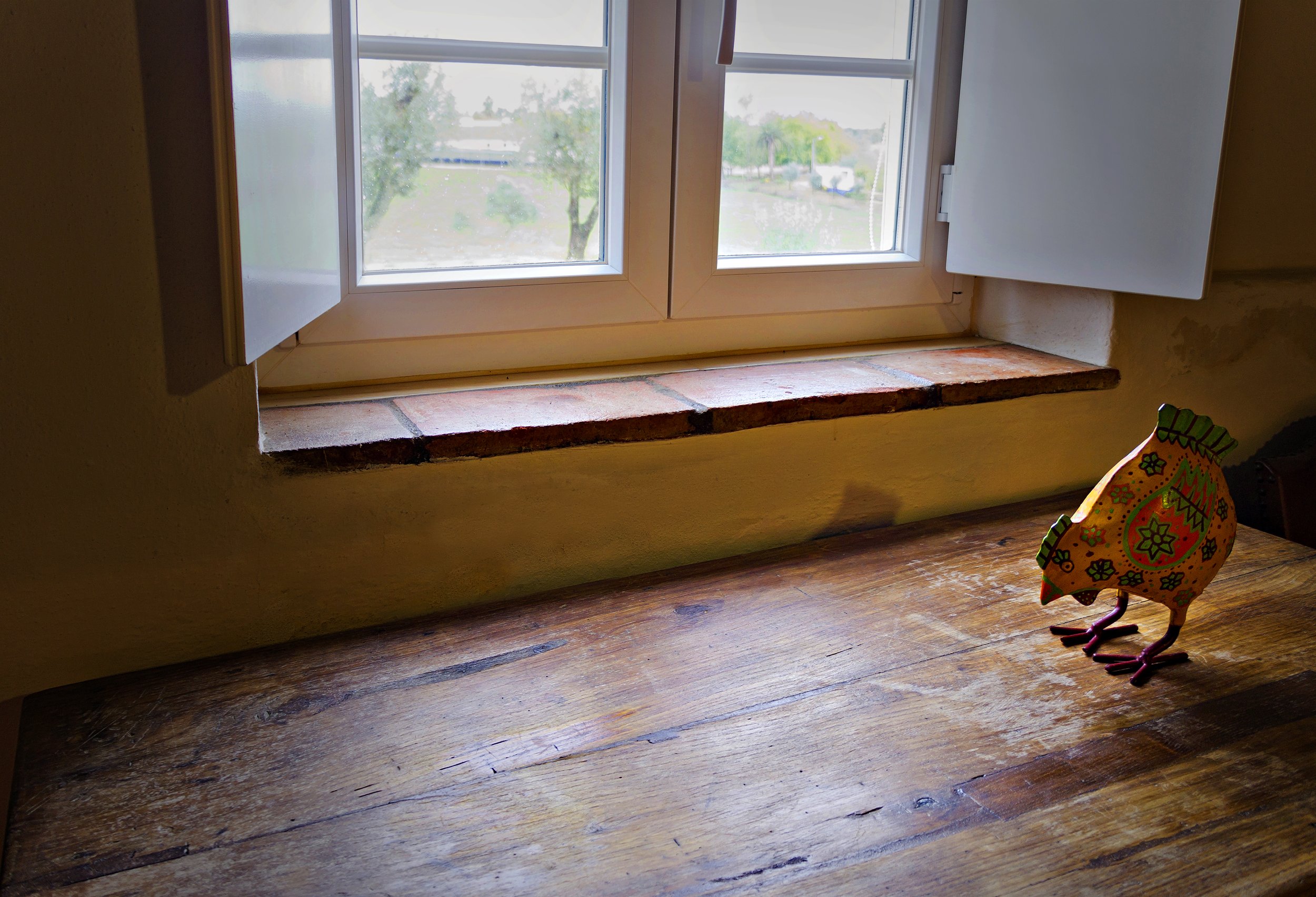 Wooden table in front of a window with white shutters, decorated with a colorful rooster sculpture.