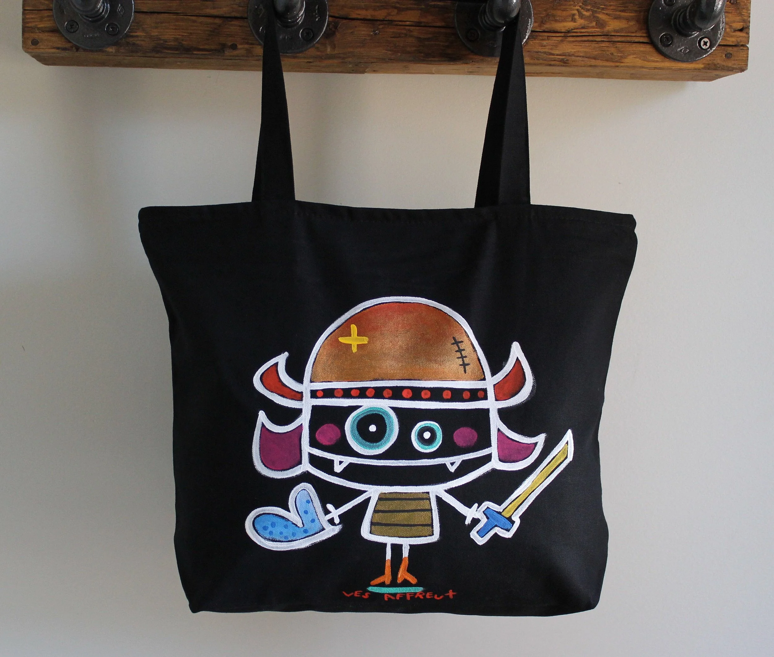 Un sac en tissu noir avec un dessin coloré d'une créature fantastique, tenant une épée et portant un chapeau, avec le texte en rouge "LES AFFEUX" en bas.