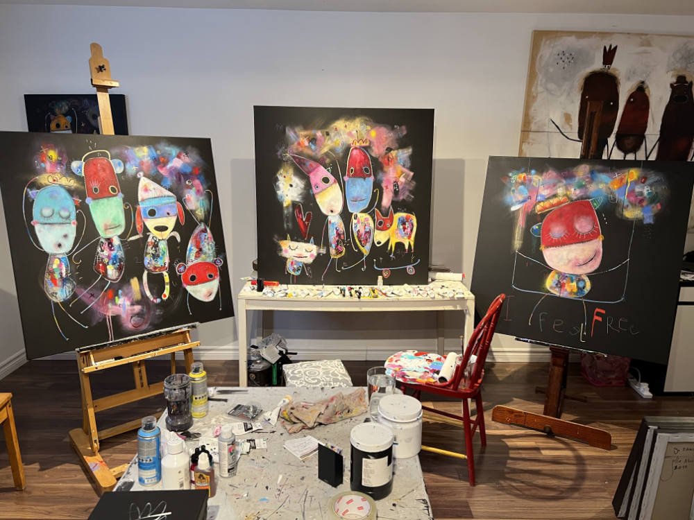 Trois toiles d'art contemporain coloré dans un style abstrait, représentant des personnages et des éléments fantaisistes, dans un atelier de peinture.