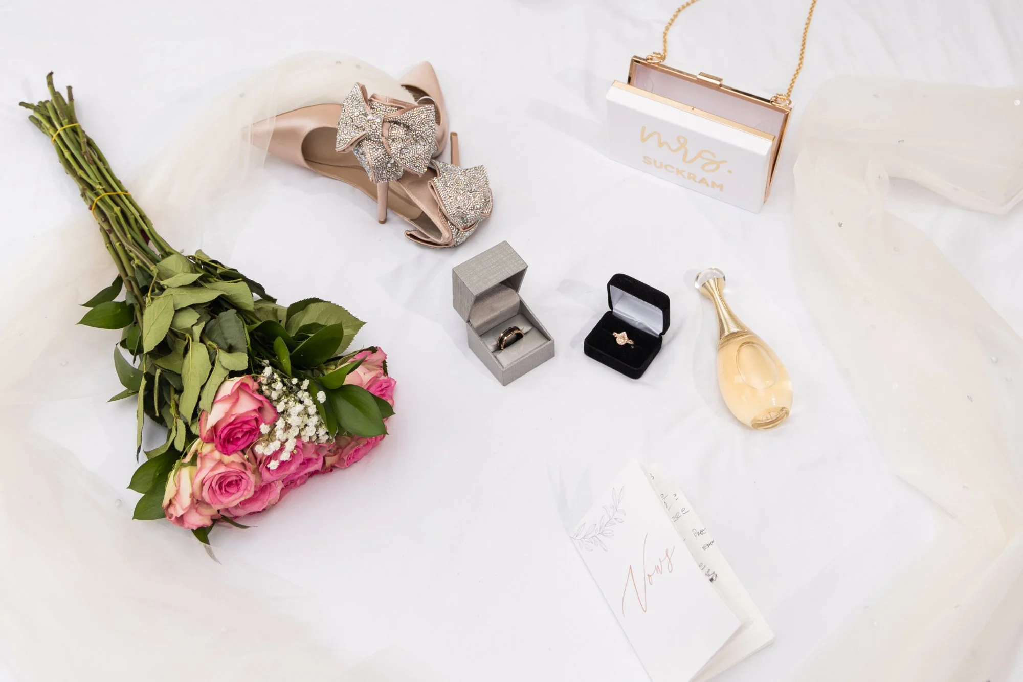 A pink bouquet of roses and white baby's breath flowers placed on a white surface, with a pair of high-heeled shoes adorned with glittery bows, a necklace in a white and gold box, two image rings in black and silver boxes, a gold-colored perfume bottle, a white card, and some white fabric or paper.