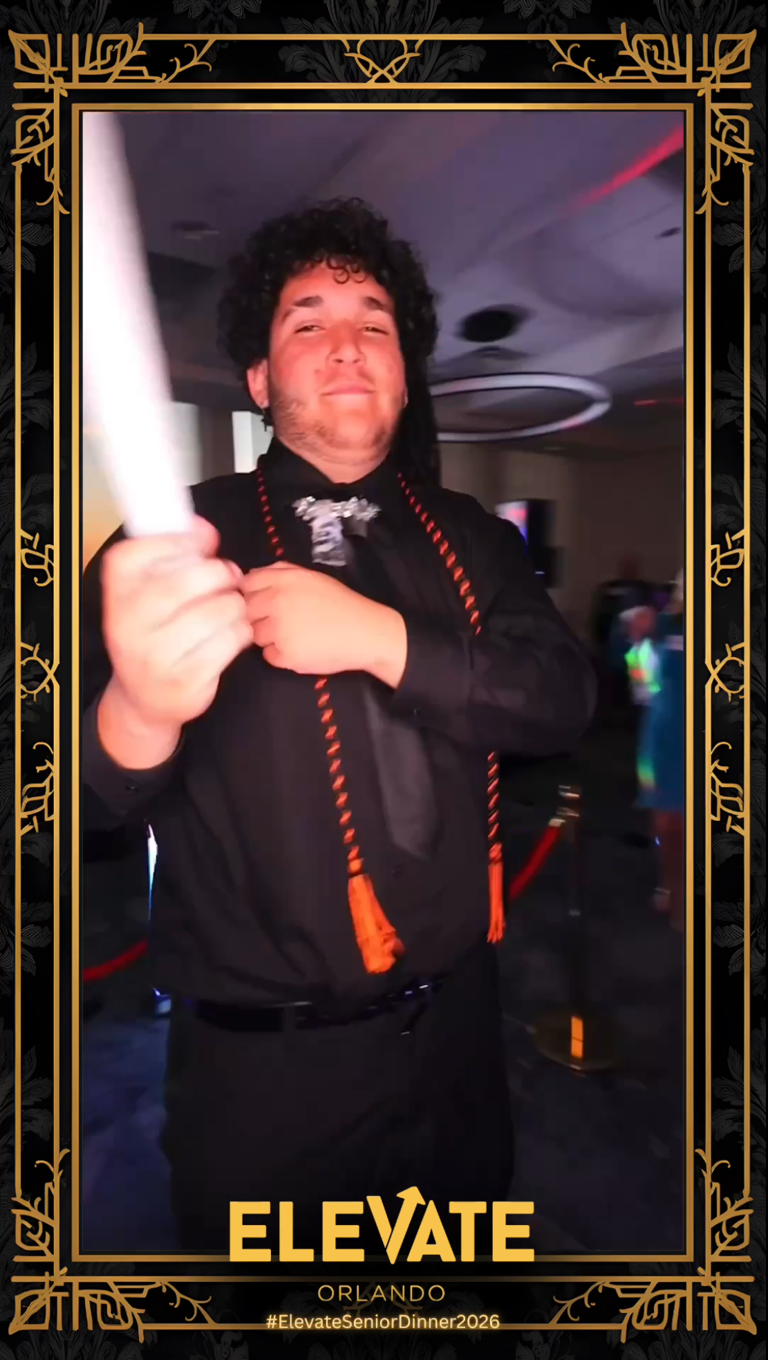 A man with curly hair dressed in a black shirt and black pants, holding a rolled-up certificate or diploma, smiling at a party or celebration event with a decorative black and gold border, and the words "ELEVATE ORLANDO" and hashtag "#ElevateSeniorDinner2026" at the bottom.
