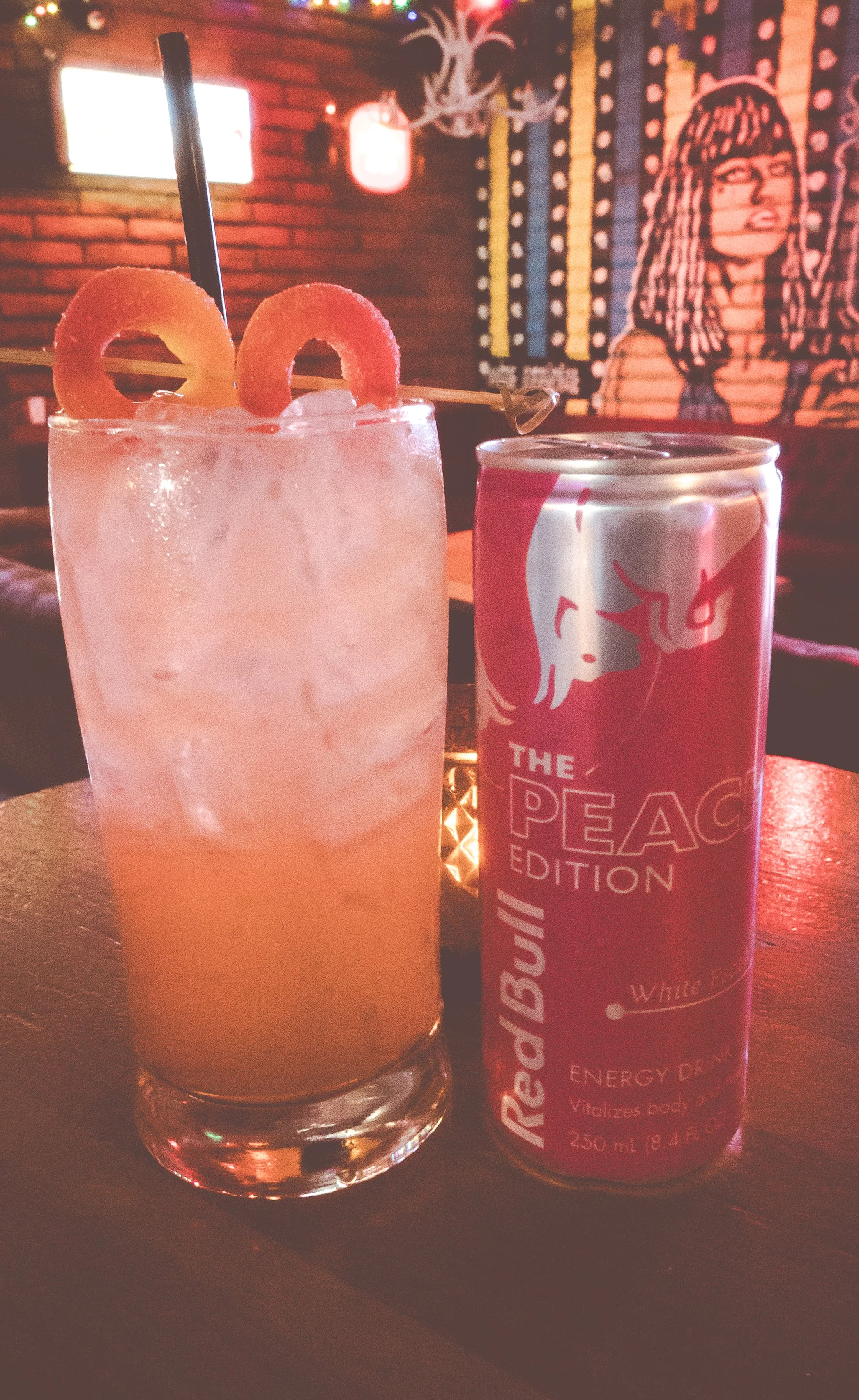 A glass of pink energy drink with gummy worms and a black straw, next to a can of Red Bull energy drink, set on a wooden table inside a bar or cafe with artwork and neon signs in the background.