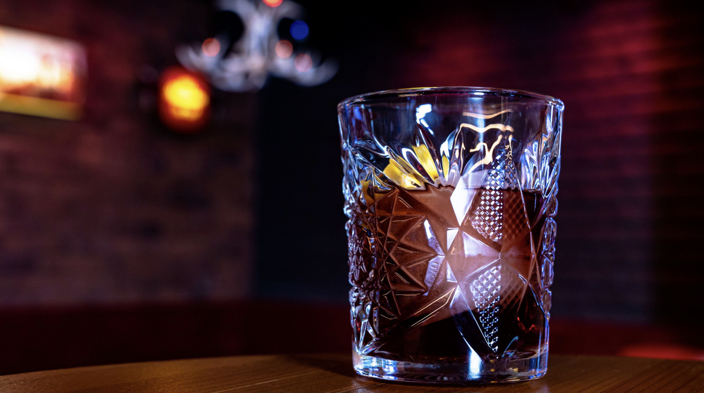A glass filled with a dark amber beverage, placed on a wooden surface, with a blurred background of colorful lights at Lucky's Indoor Outdoor bar in Phoenix AZ