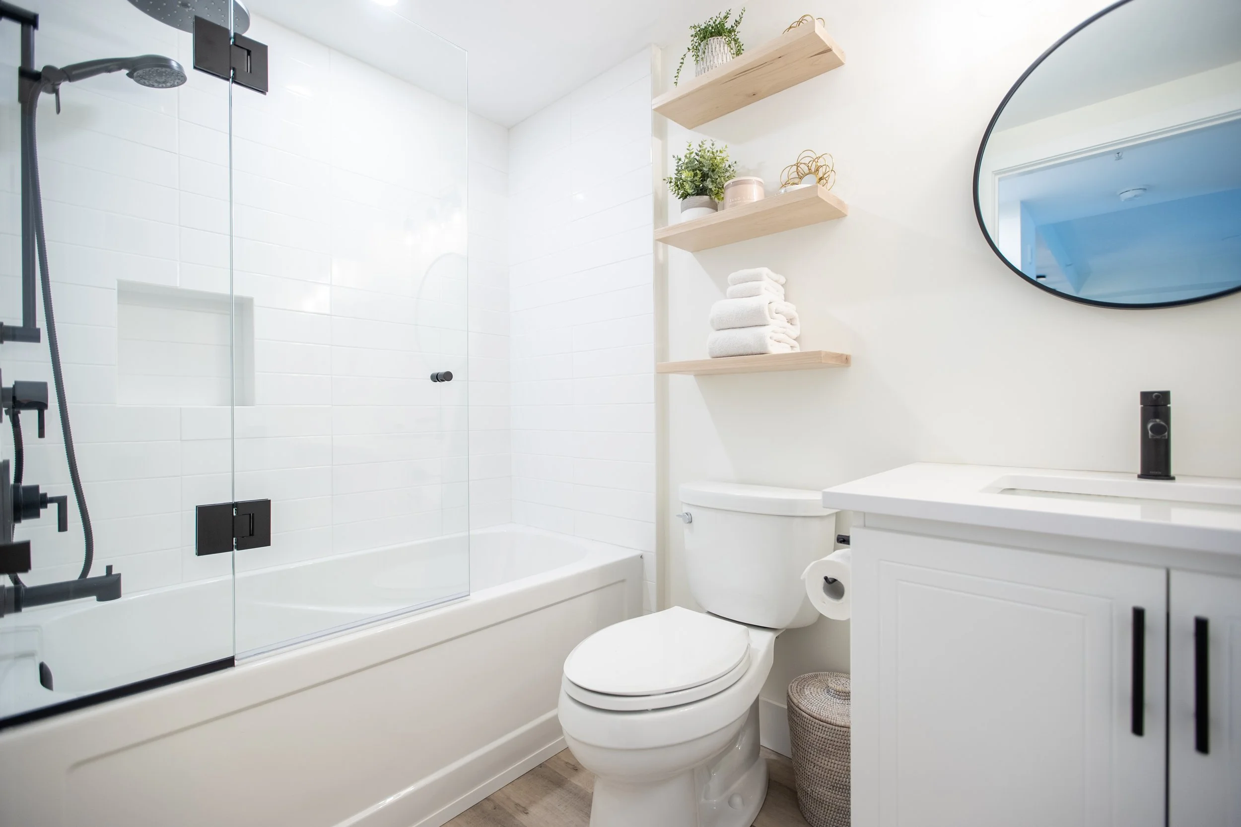 Bathroom with a white bathtub, custom glass shower door, white toilet, custom wooden shelves, round mirror, white vanity with a black faucet, and light-colored Luxury Vinyl Plank (LVP) flooring.