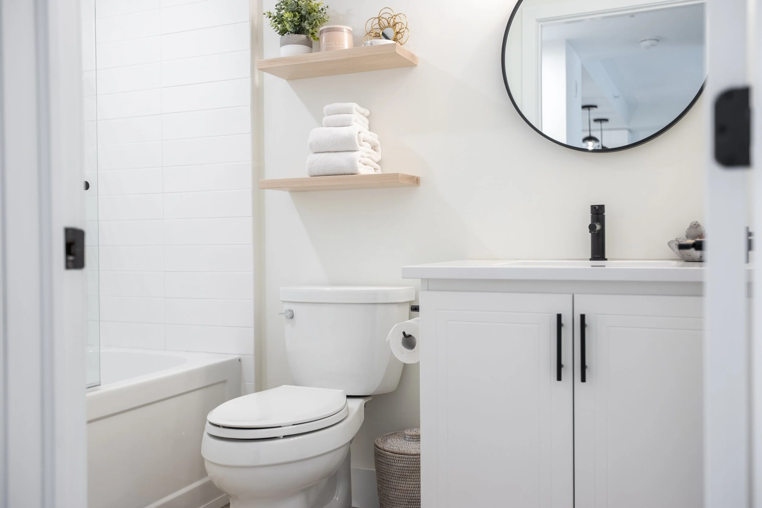 Bathroom with white walls, a shower with white tiles, white toilet, a white vanity with black handles, a round mirror, black faucet, and wooden shelves holding towels and decor.