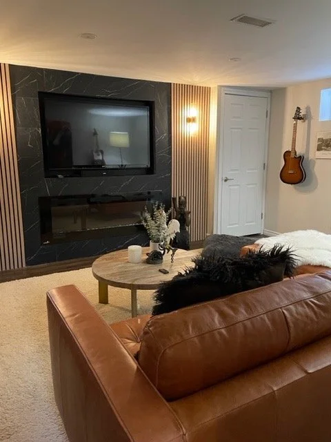 Living room with wall-mounted TV, fireplace, round coffee table with decorative items, brown leather couch with black pillow, white blanket, and a guitar on the wall.