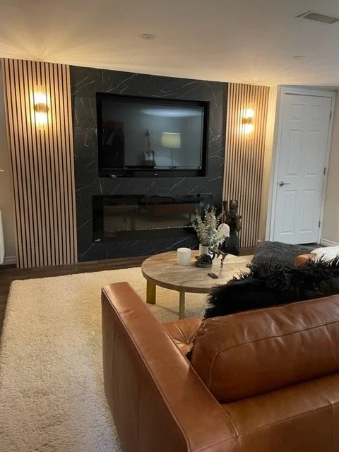 Living room with a wall-mounted TV surrounded by black marble and wooden paneling, a round wooden coffee table with decorative items, a brown leather sofa with black pillows, a beige rug, and a closed white door.
