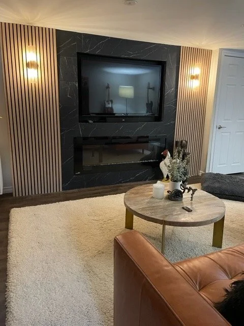 Living room with a black marble fireplace, beige walls with vertical wooden paneling, a beige rug, a round coffee table with decorative items, and a leather chair.