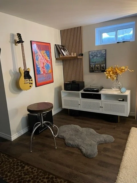 Room with a wall-mounted yellow guitar, a framed colorful artwork, a small white media console with a vase of yellow flowers, a stool, and a window showing blue sky.