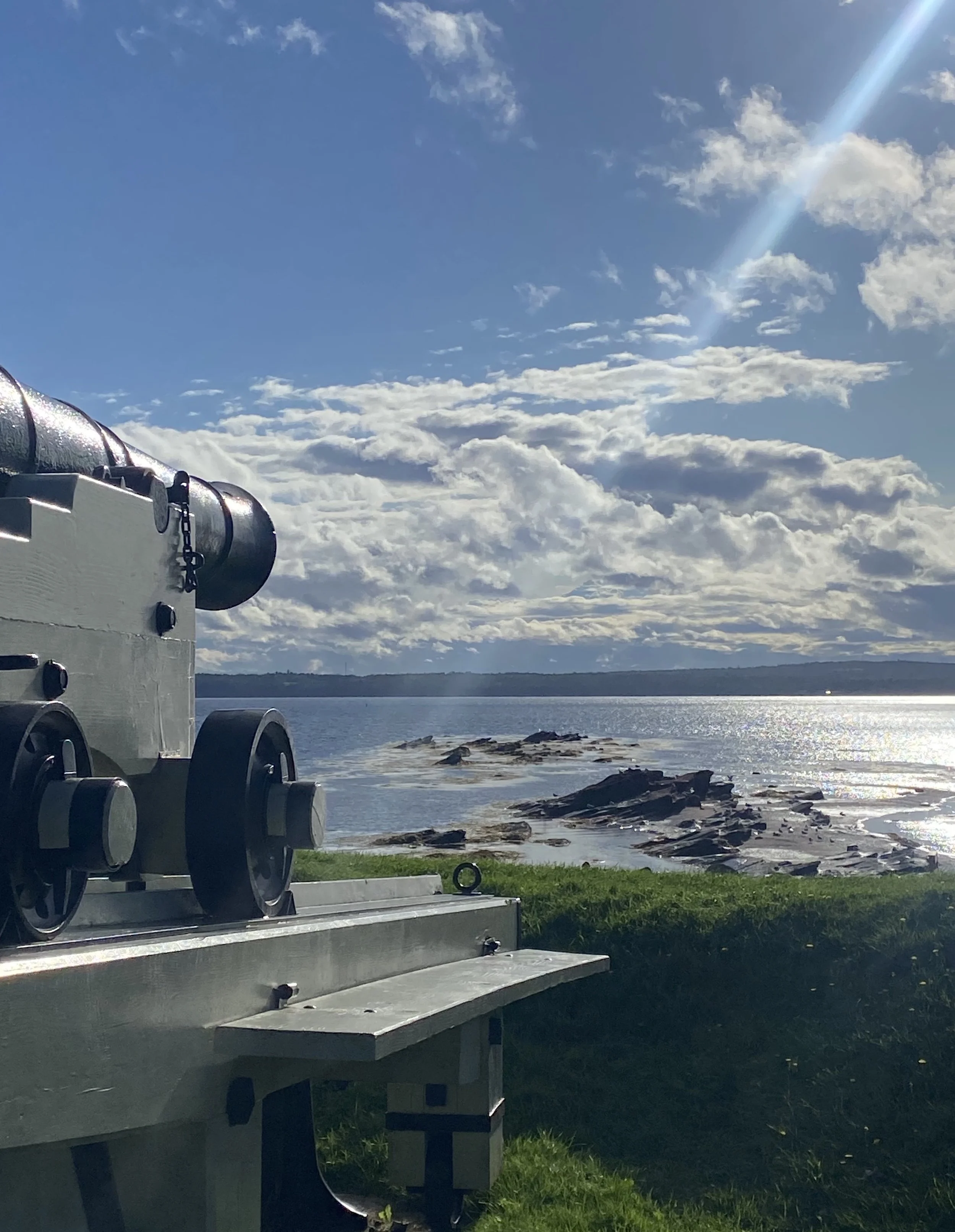 A large antique cannon on a grassy lawn overlooking a body of water with rocks, with a partly cloudy sky and sunlight shining through clouds.
