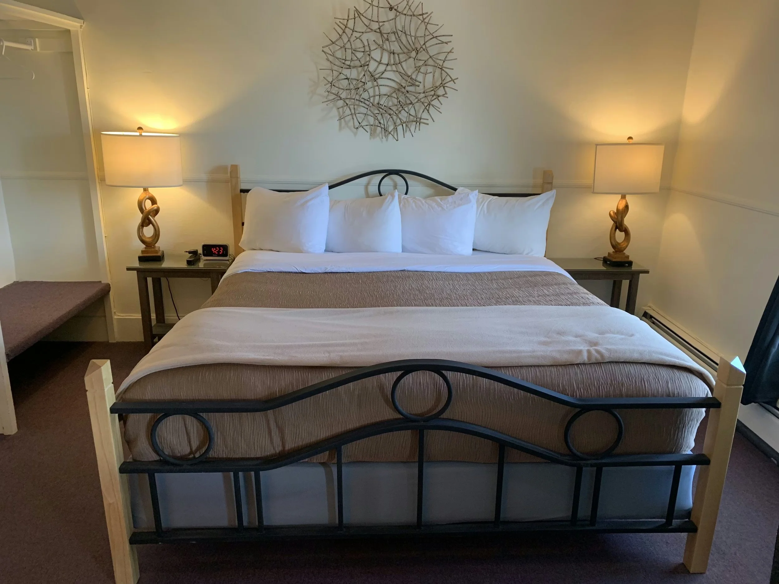 A neatly made bed with six white pillows, a beige bedspread, and a white blanket. Two matching lamps sit on bedside tables on either side of the bed. A decorative wall hanging is centered above the headboard. There is a digital clock showing 4:23 on the left bedside table and a small black device. The room has beige walls and carpet.
