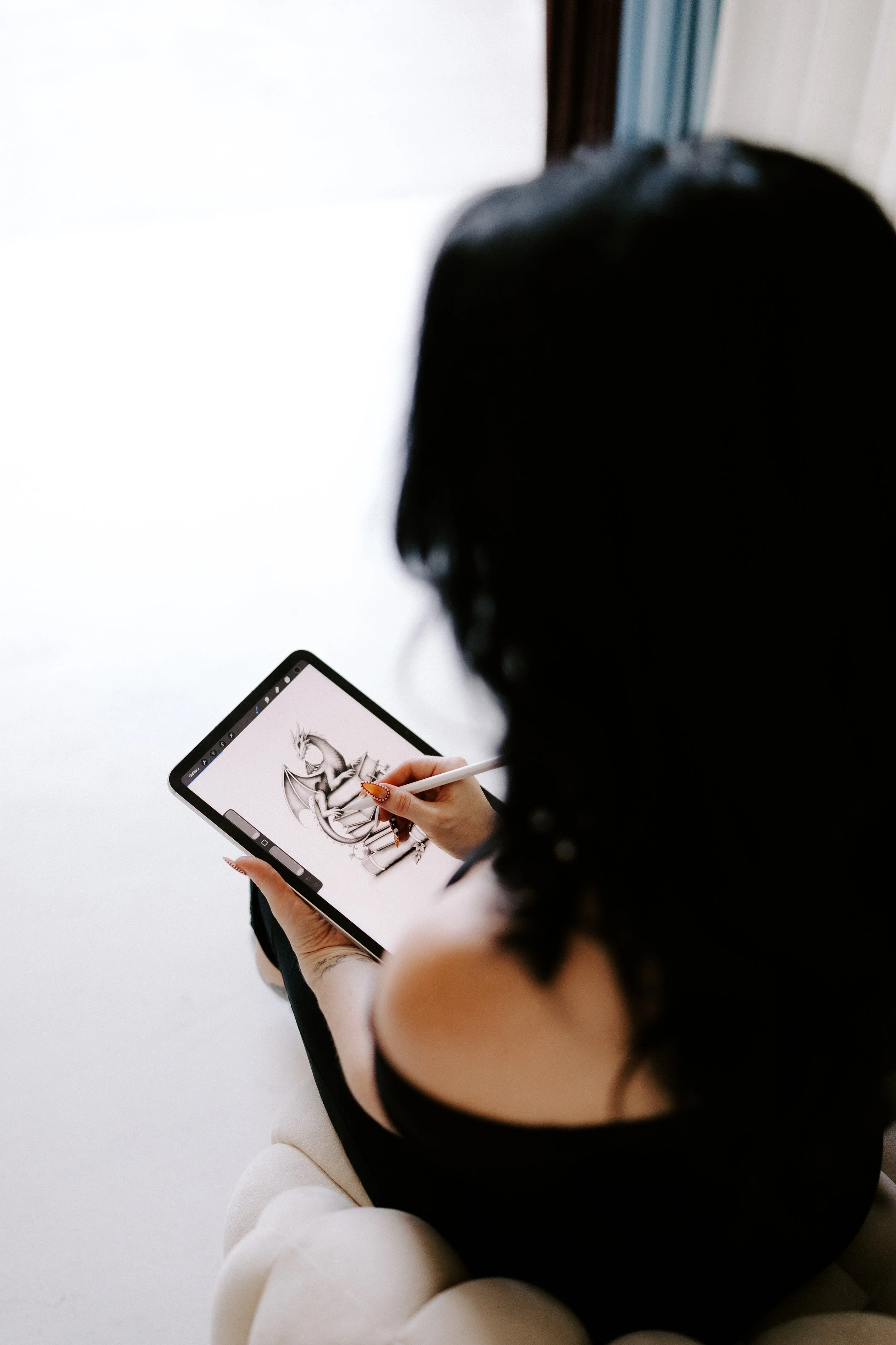 Person with dark hair drawing a detailed sketch of a dragon on a tablet with a stylus, seated on a white surface.