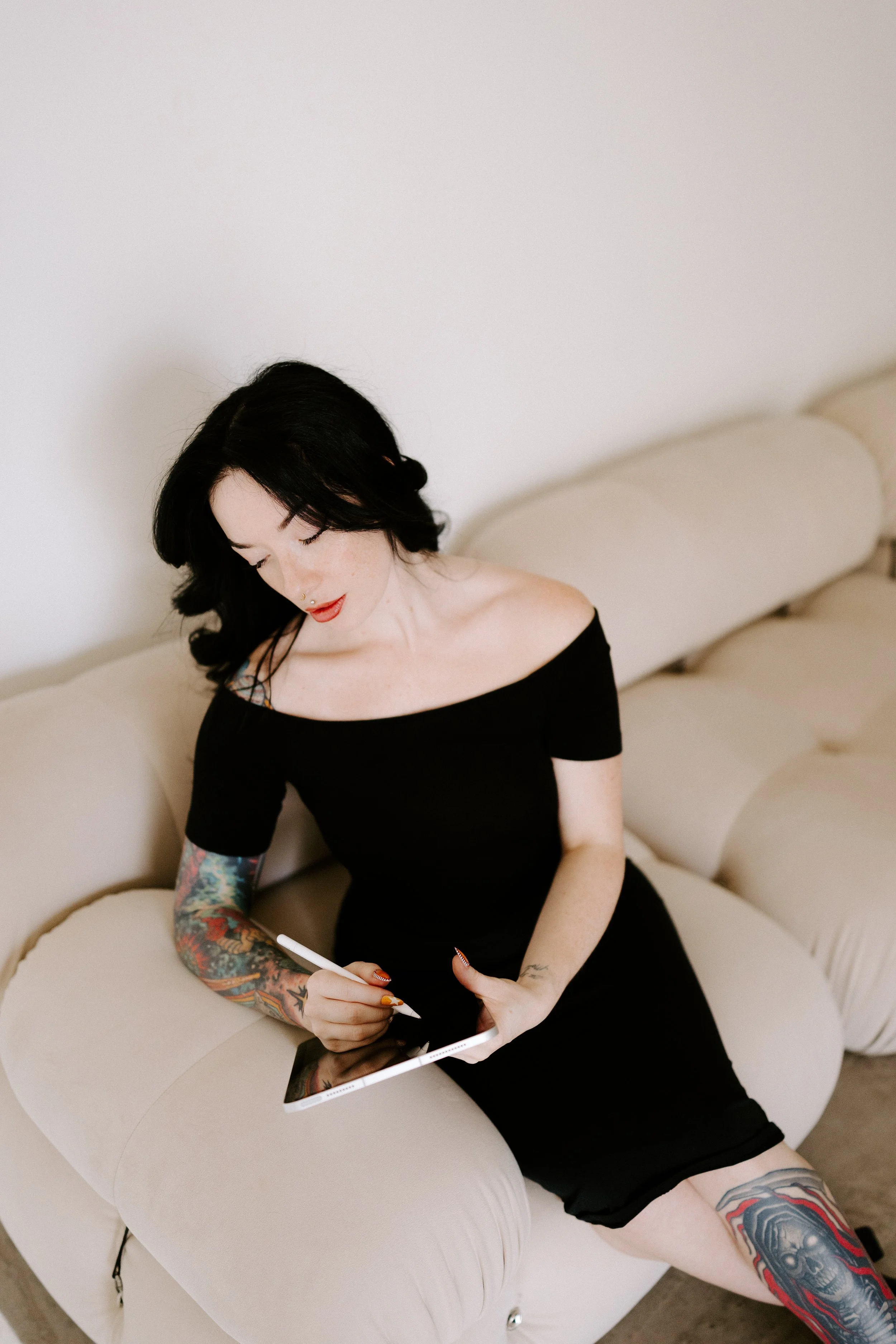 Woman with black hair and tattoos, sitting on a beige curved sofa, using a stylus on a tablet.