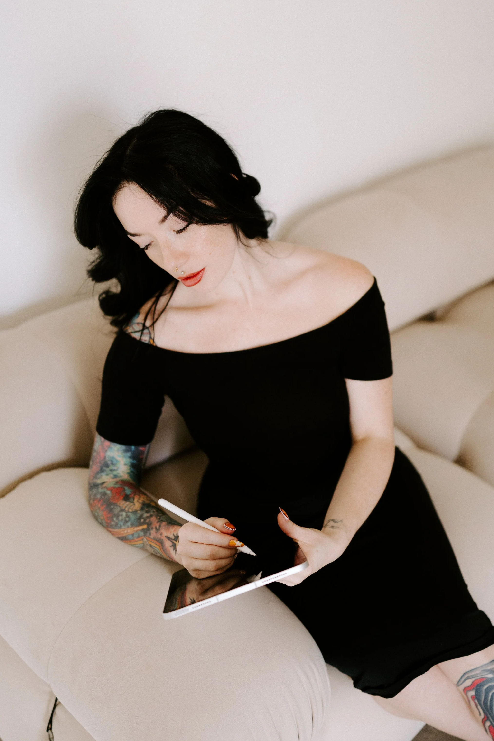 A woman with black hair, red lipstick, and tattooed arm sitting on a beige couch, using a tablet with a stylus.