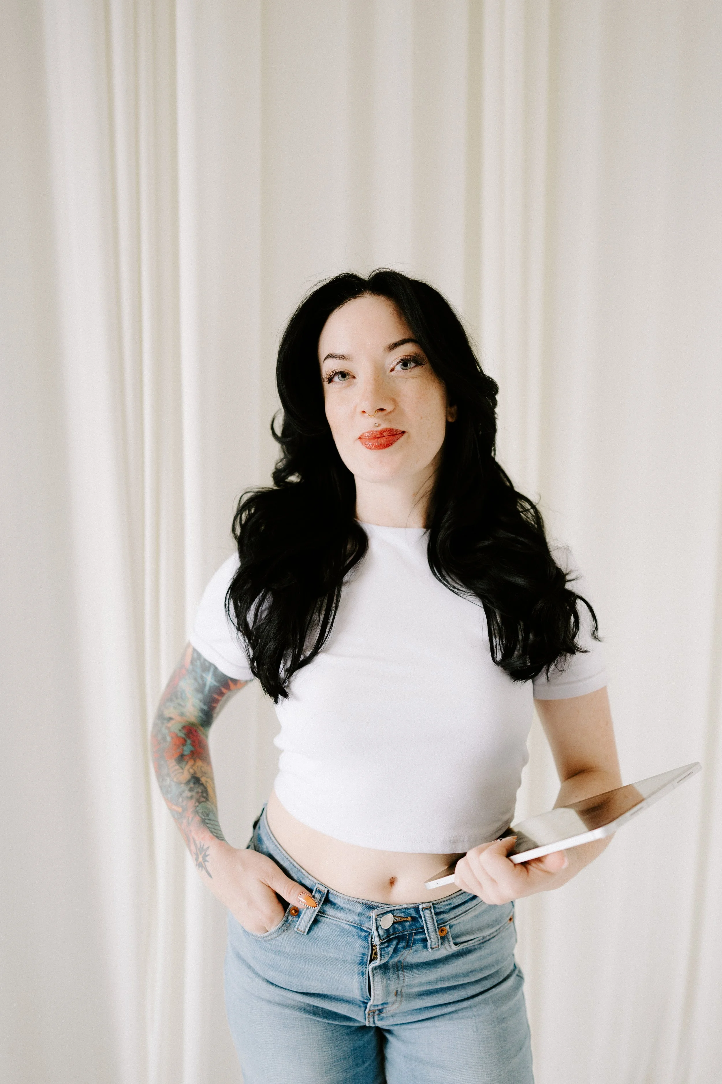 A woman with long black hair, tattoos on her left arm, wearing a white crop top and blue jeans, holding a tablet in her left hand, standing in front of a light-colored curtain.