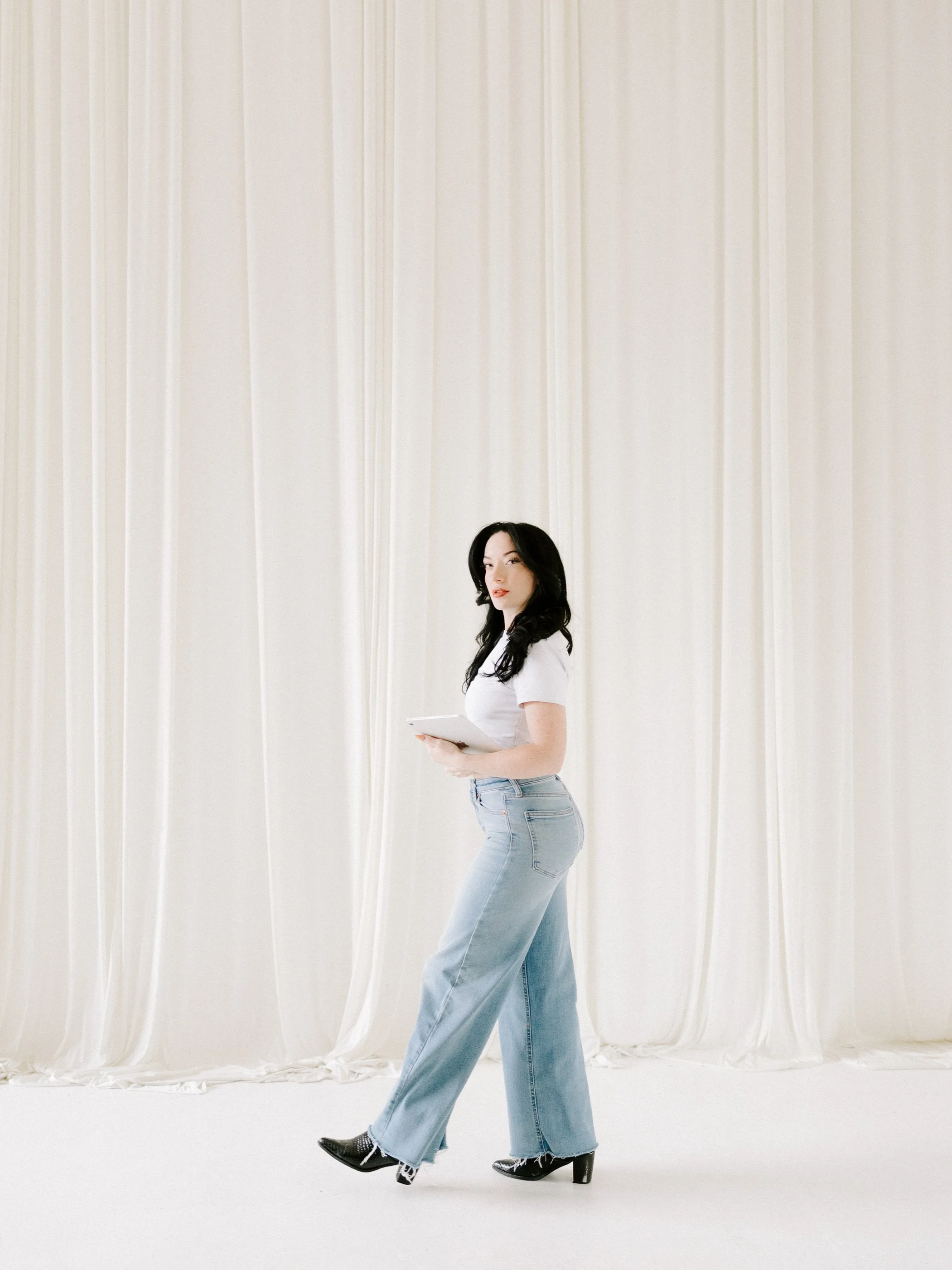 A woman with long black hair wearing a white t-shirt, light blue jeans, and black high-heeled shoes, standing against a cream-colored curtain backdrop, holding a white tablet.