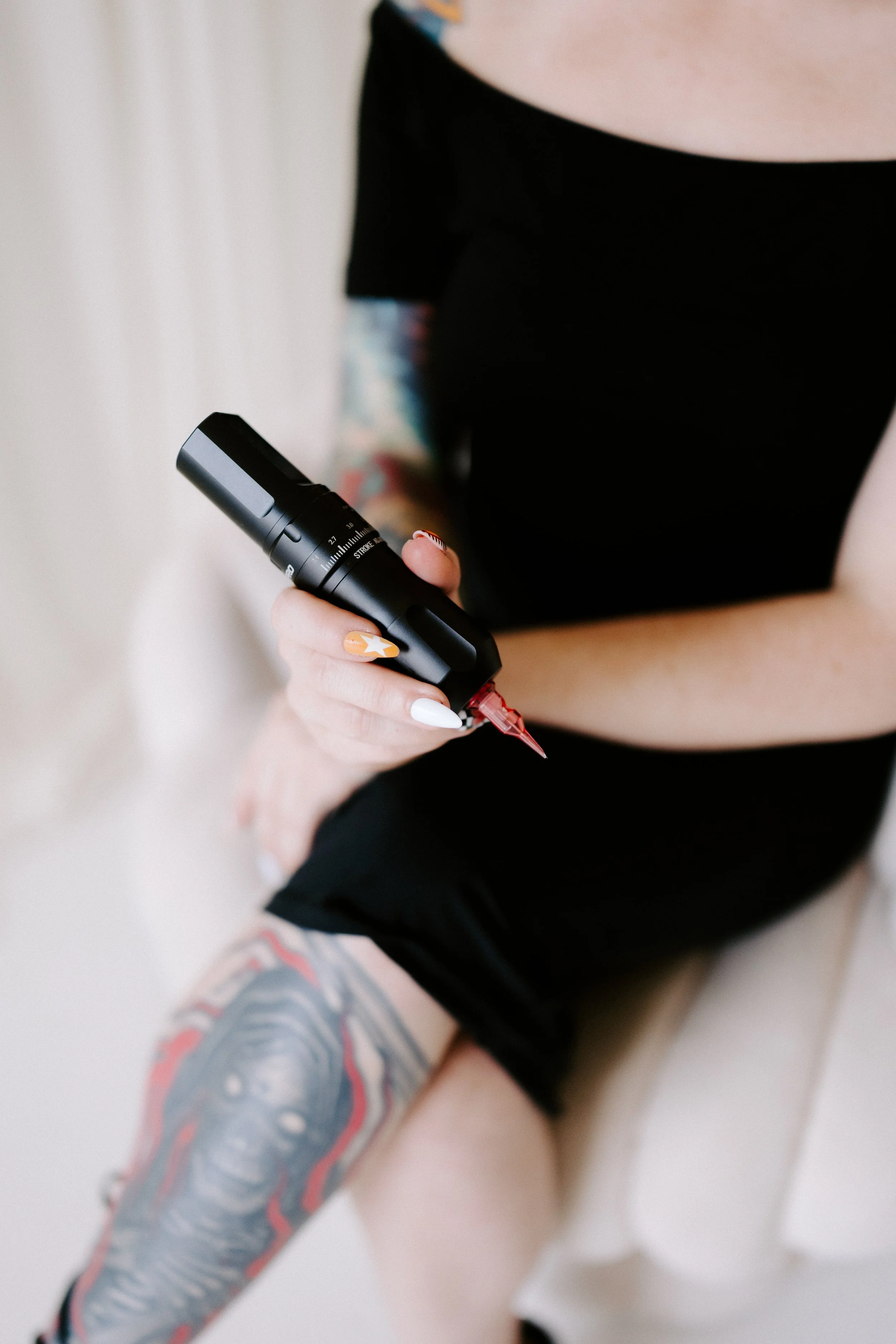 Close-up of a person holding a black tattoo machine, with visible tattooed arm and leg, in a tattoo studio setting.