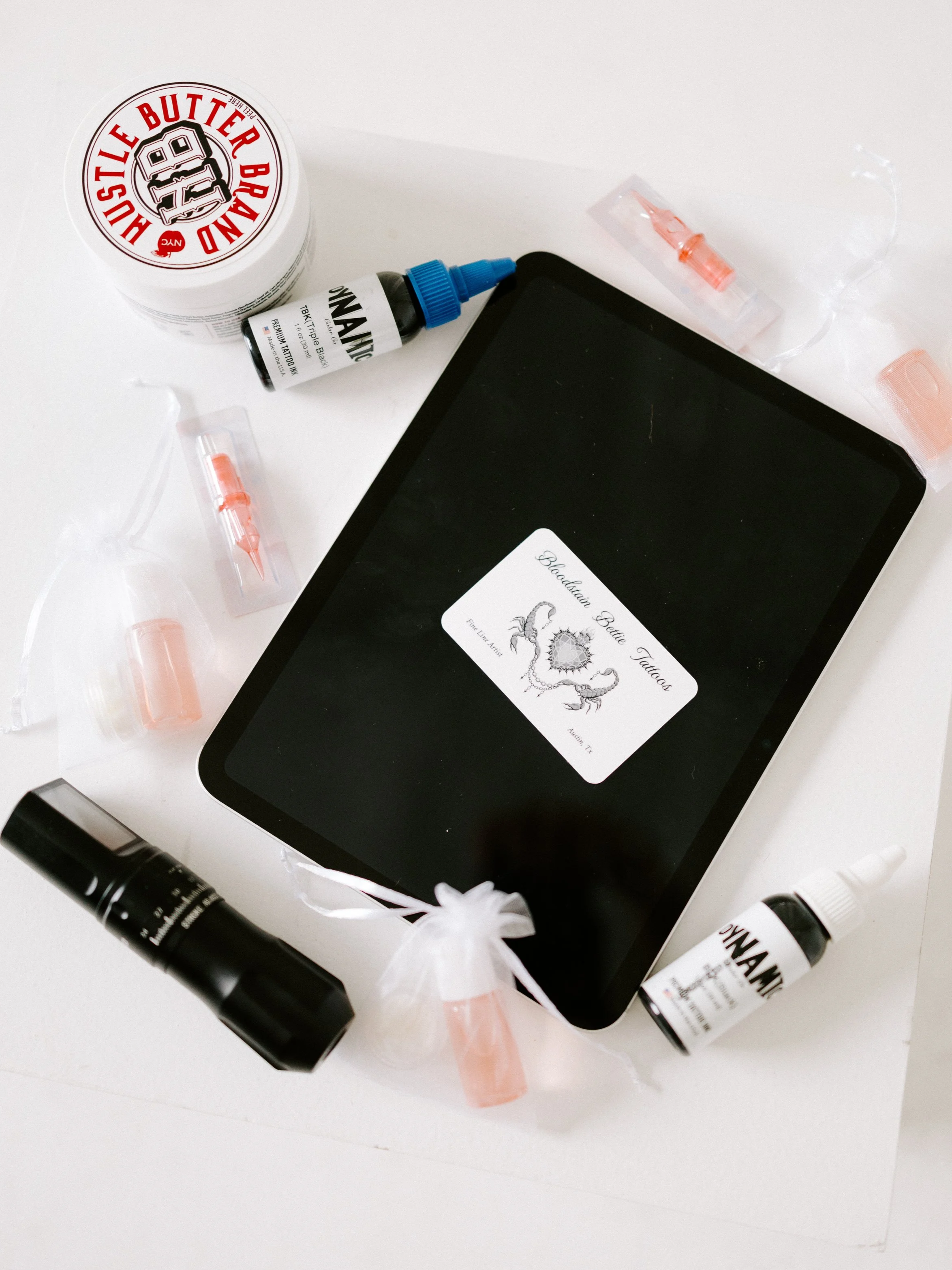 A tablet with a tattoo design titled 'Bloodrain Bettie Tattoos' surrounded by tattoo ink bottles, tattoo needles, and tattoo ink tubes on a white surface.